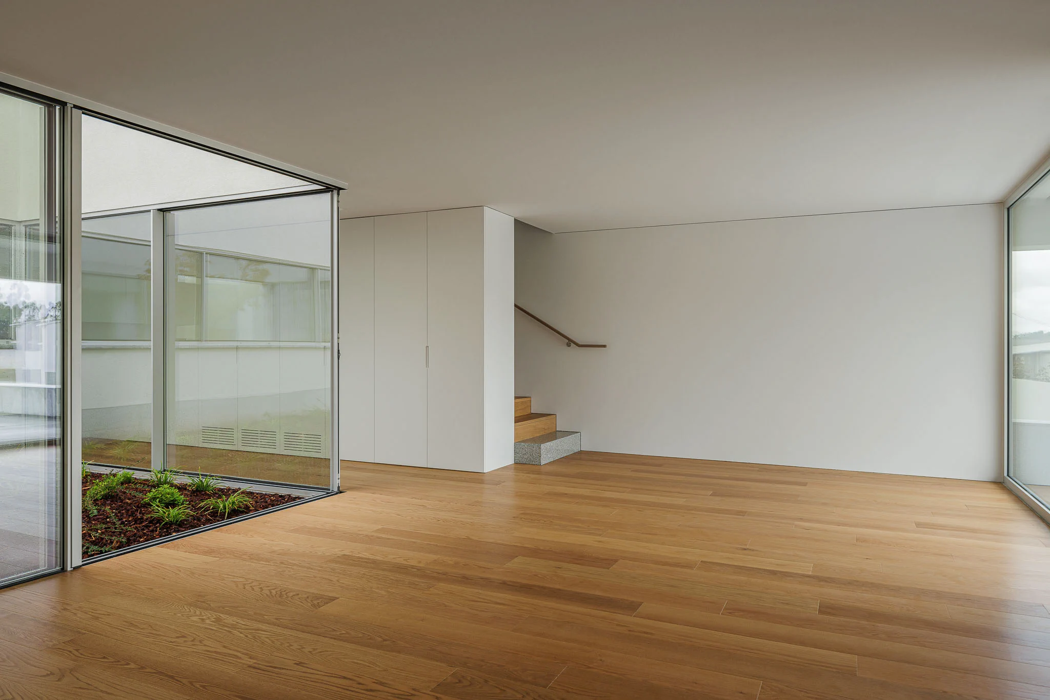 Sala ampla com pavimento em madeira, paredes brancas, grandes janelas envidraçadas e escada ao fundo. — Casa em Santa Marinha arquitetura Helder da Rocha Arquitectos em Lousada