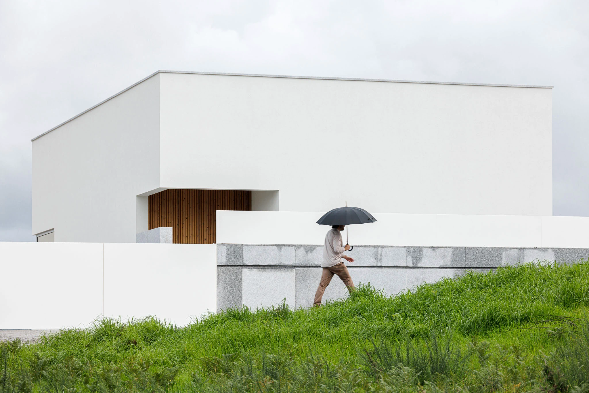 Homem com guarda-chuva a caminhar junto à casa branca, com base em granito e vegetação rasteira. — Casa em Santa Marinha arquitetura Helder da Rocha Arquitectos em Lousada