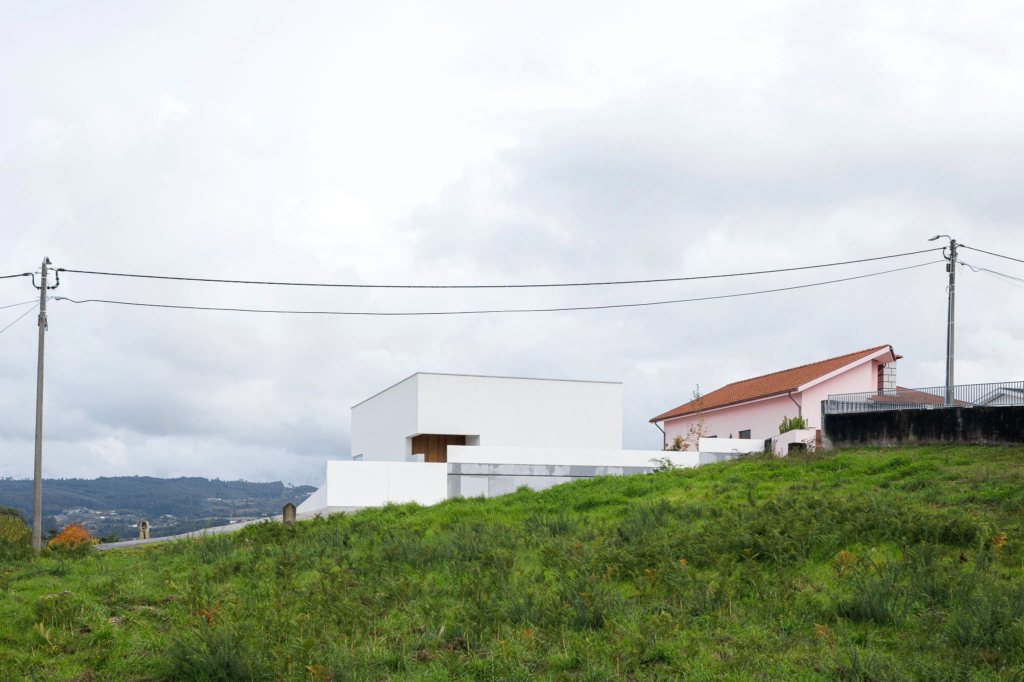 Vista da casa branca no topo de uma colina, com casa rosa vizinha e paisagem rural ao fundo. — Casa em Santa Marinha arquitetura Helder da Rocha Arquitectos em Lousada