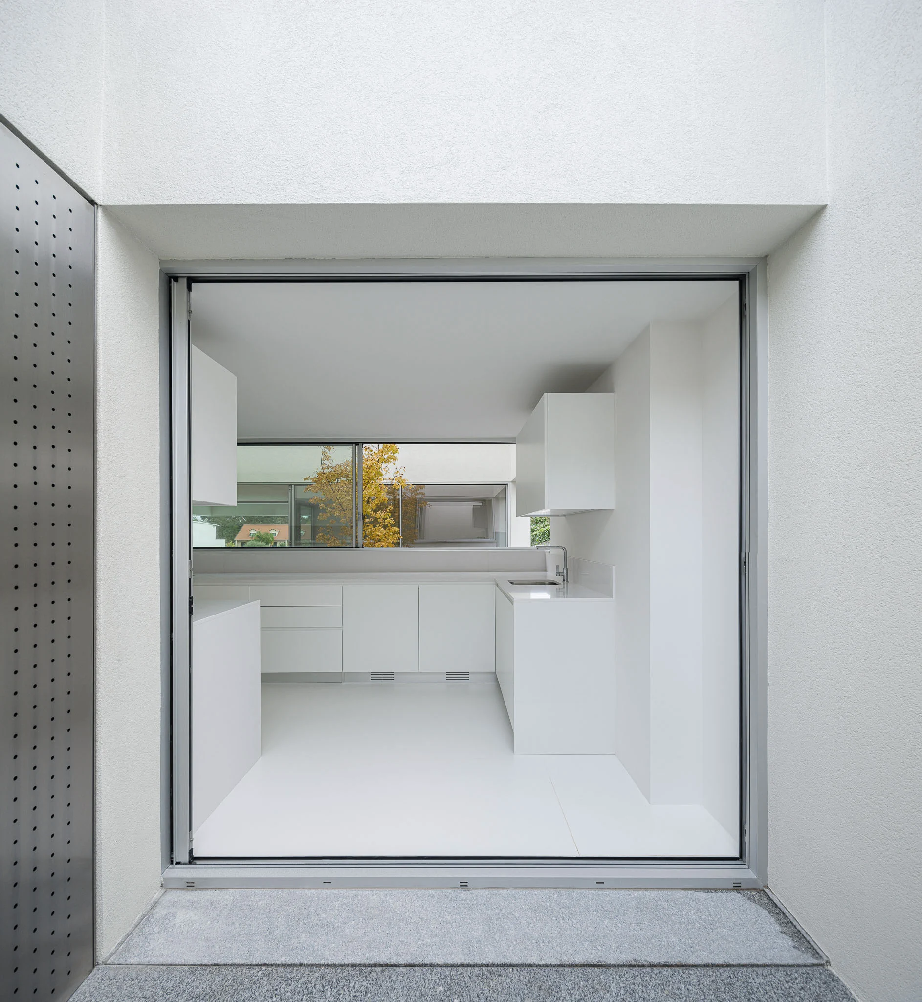 Vista da cozinha branca através de porta metálica perfurada, com pátio interior visível ao fundo. — Casa em Santa Marinha arquitetura Helder da Rocha Arquitectos em Lousada