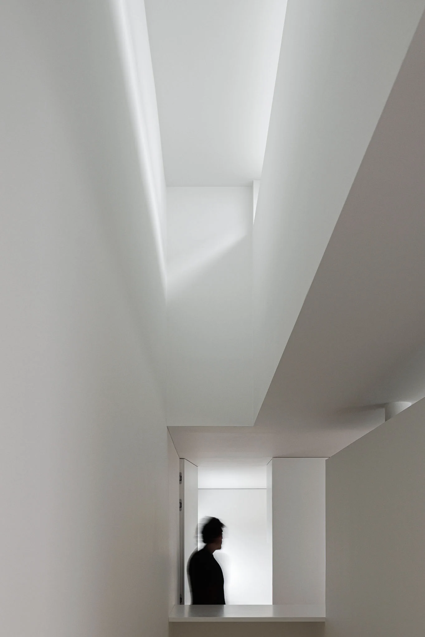 Espaço interior com paredes brancas, iluminação zenital e pessoa desfocada ao fundo. — Casa em Santa Marinha arquitetura Helder da Rocha Arquitectos em Lousada