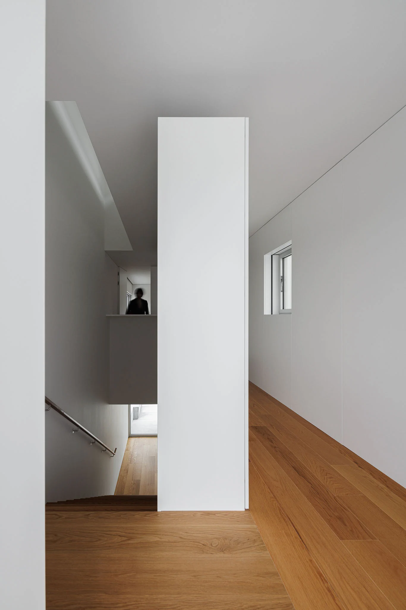 Corredor com pavimento em madeira, paredes brancas, escada e pessoa ao fundo. — Casa em Santa Marinha arquitetura Helder da Rocha Arquitectos em Lousada