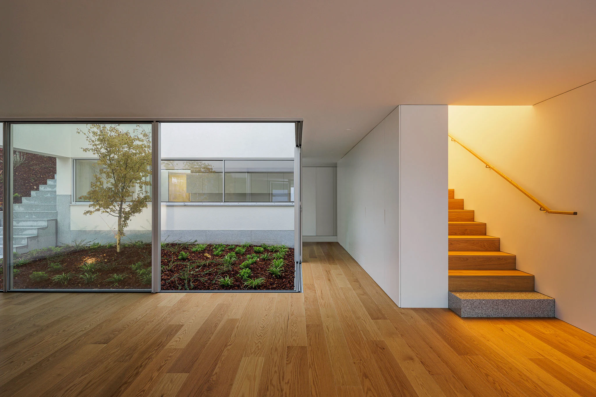 Sala com vista para pátio interior e escada interior iluminada, com pavimento em madeira. — Casa em Santa Marinha arquitetura Helder da Rocha Arquitectos em Lousada