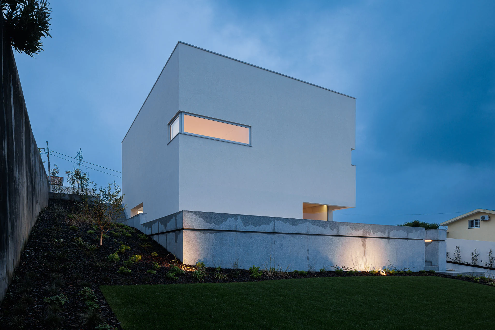 Vista noturna da casa branca com base em granito, iluminação exterior e jardim relvado. — Casa em Santa Marinha arquitetura Helder da Rocha Arquitectos em Lousada