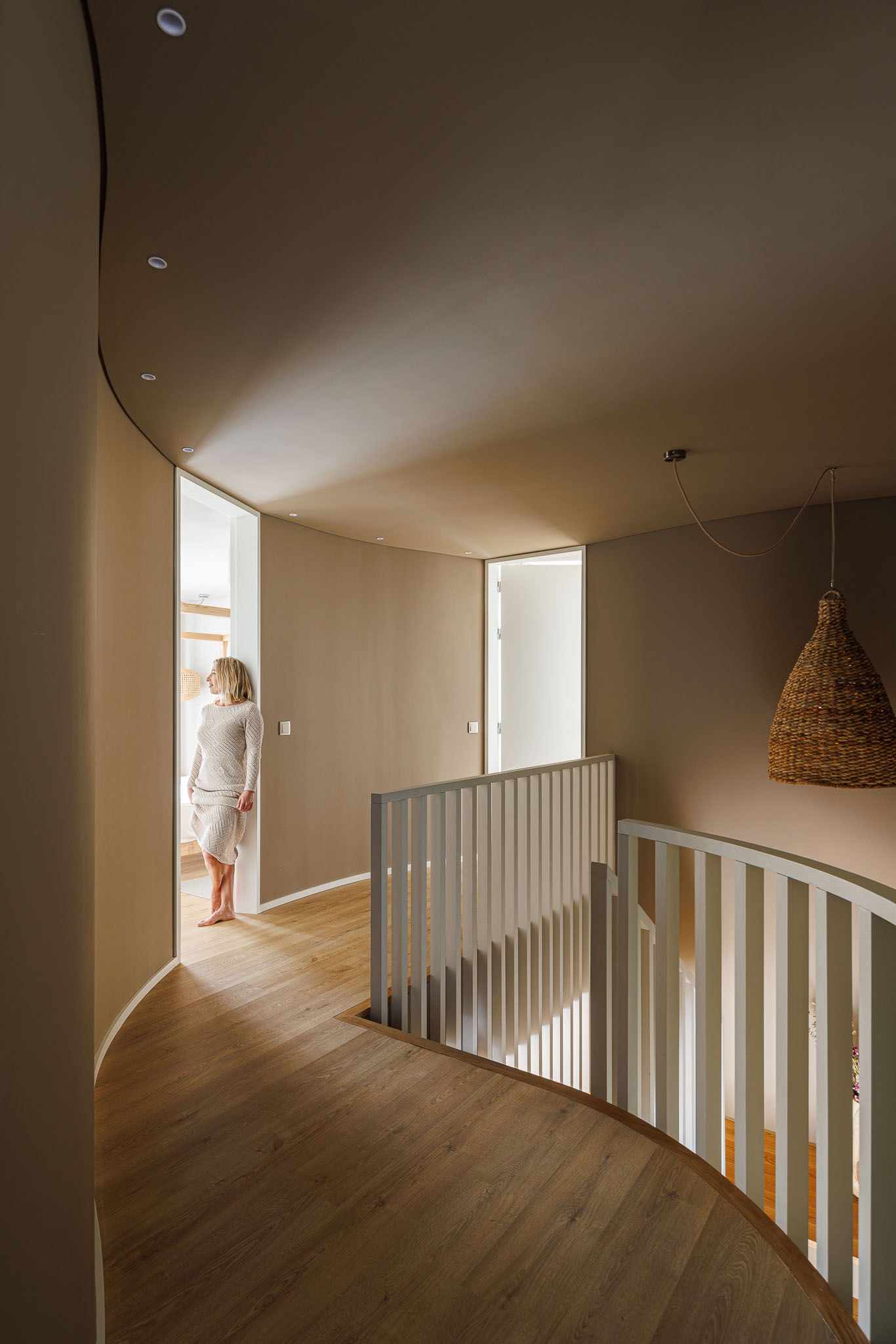 Corredor: Pavimento em madeira, paredes curvas em tons terra e candeeiro de vime suspenso. — Casa Pátios de Pétalas arquitetura Sandra Micaela Casinha Atelier em Gondomar