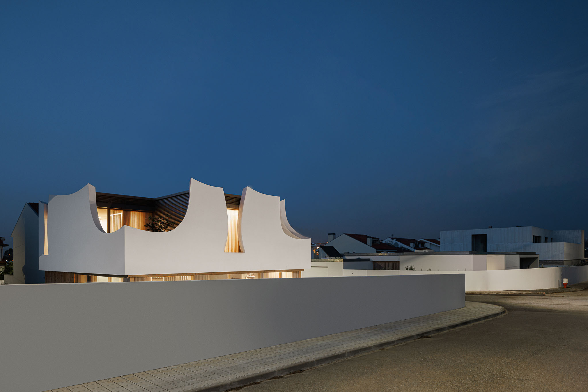 Exterior: Vista noturna da casa com detalhes curvos e iluminação interior. — Casa Pátios de Pétalas arquitetura Sandra Micaela Casinha Atelier em Gondomar