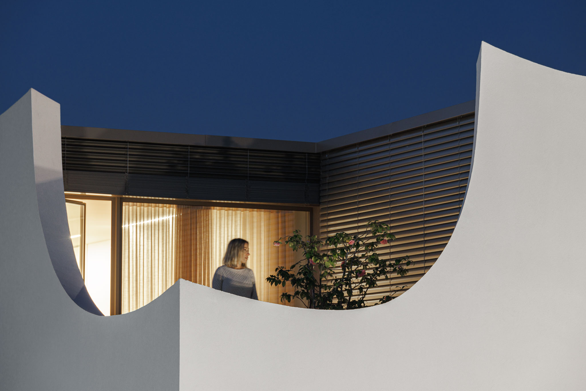 Exterior: Detalhe da varanda curva branca com ripas horizontais e vegetação. — Casa Pátios de Pétalas arquitetura Sandra Micaela Casinha Atelier em Gondomar