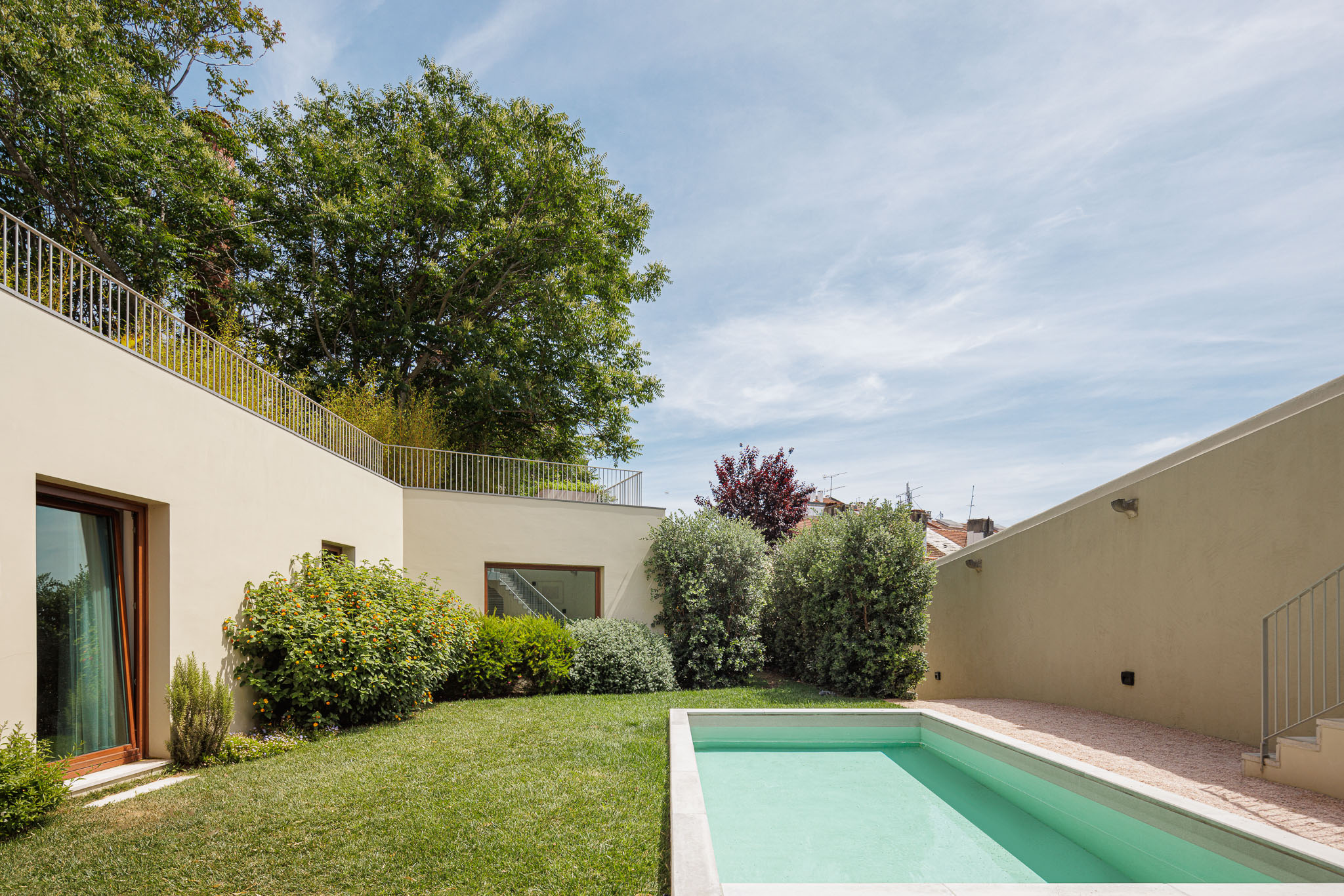 Exterior: Piscina retangular em betão, relvado e vegetação densa. Fachada da casa em tons neutros com janelas em madeira. — Prazeres 37 arquitetura Fragmentos em Lisboa