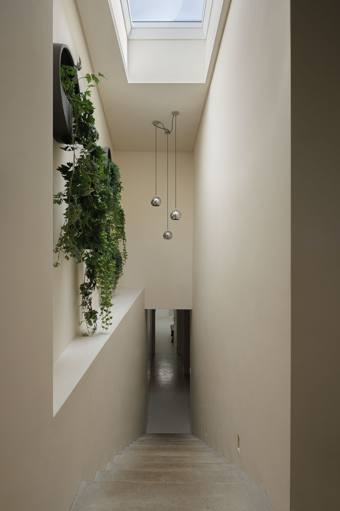 Escadas: Corredor vertical iluminado por claraboia e candeeiro suspenso. Paredes caiadas e plantas penduradas. — Prazeres 37 arquitetura Fragmentos em Lisboa
