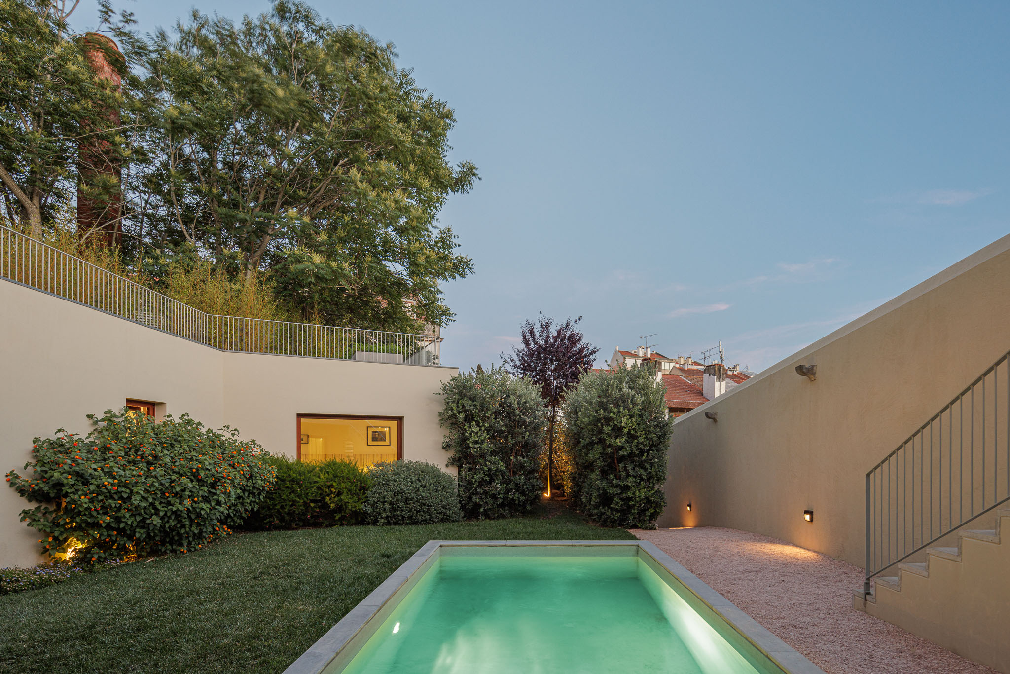 Exterior: Piscina retangular com iluminação noturna, relvado e vegetação. Fachada da casa em tons neutros. — Prazeres 37 arquitetura Fragmentos em Lisboa