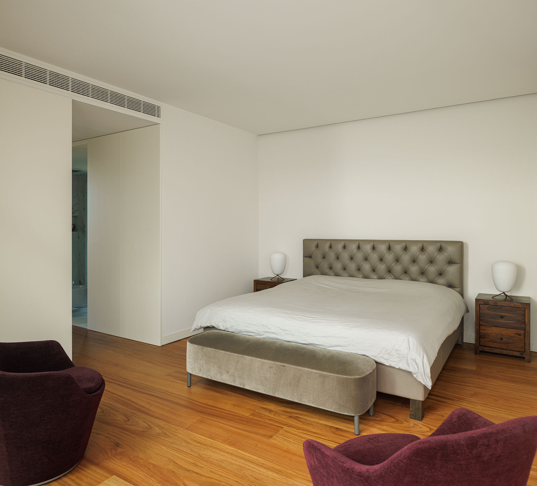 Quarto: Cama com cabeceira almofadada, mesinhas de cabeceira em madeira e cadeirões em tons de vinho. Pavimento em madeira. — CONCRETE 11 arquitetura Cage Atelier em Lisboa