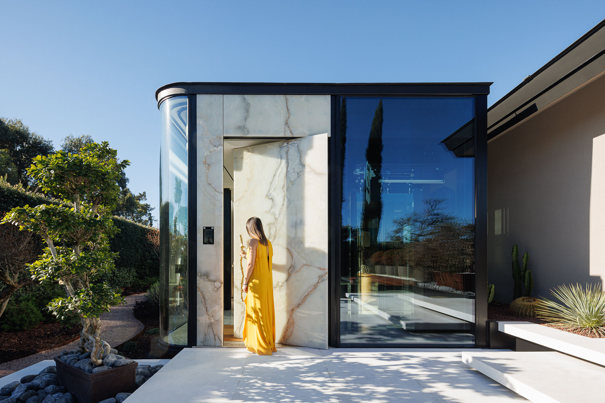 Entrada: Porta pivotante em ónix translúcido, emoldurada por vidro e metal preto, com jardim exterior. — Casa de Lavra arquitetura Ricardo Azevedo Arquitecto em Matosinhos