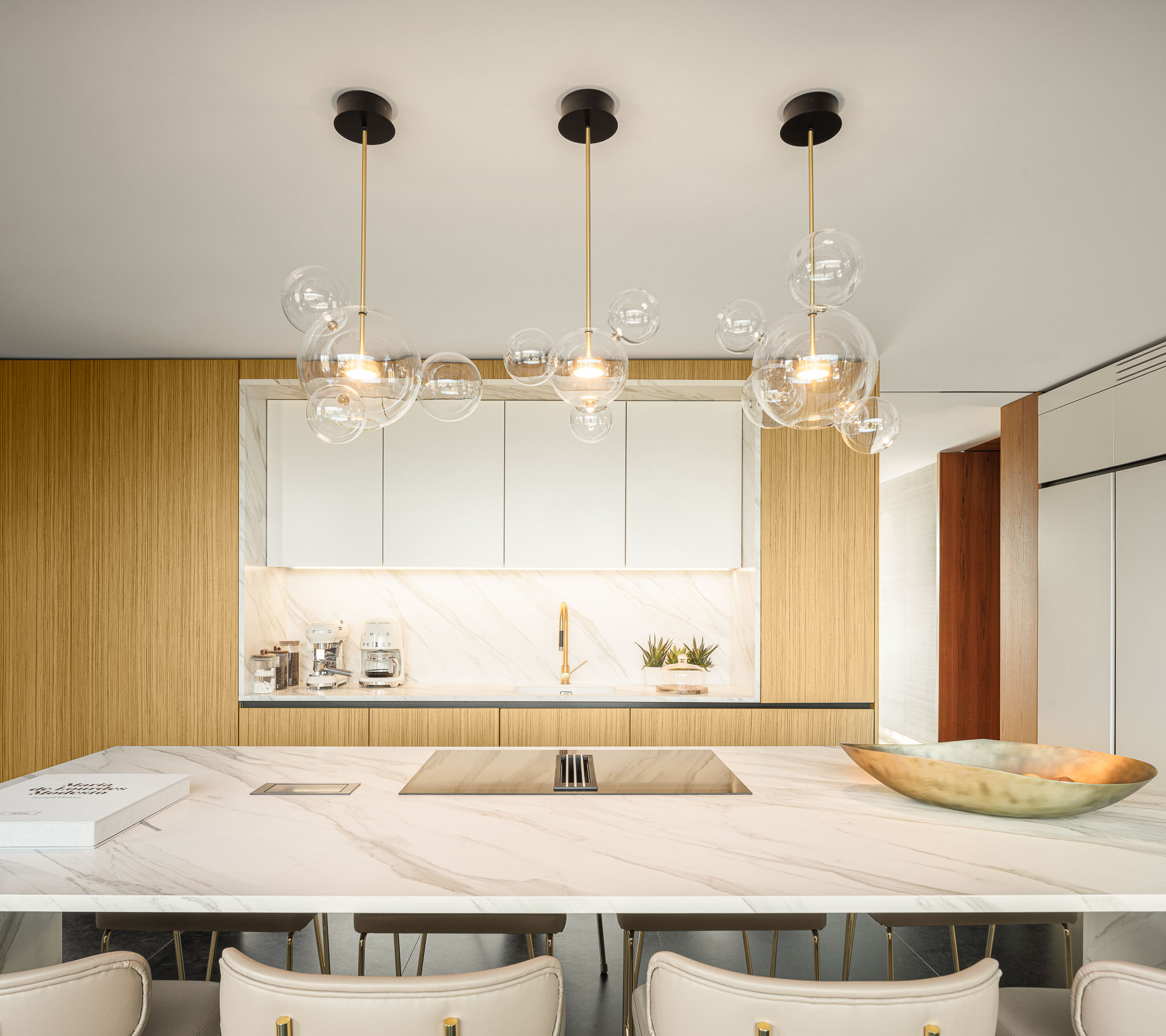 Cozinha: Ilha central em mármore com banquetas douradas, armários brancos e candeeiros suspensos de vidro. — Casa de Lavra arquitetura Ricardo Azevedo Arquitecto em Matosinhos
