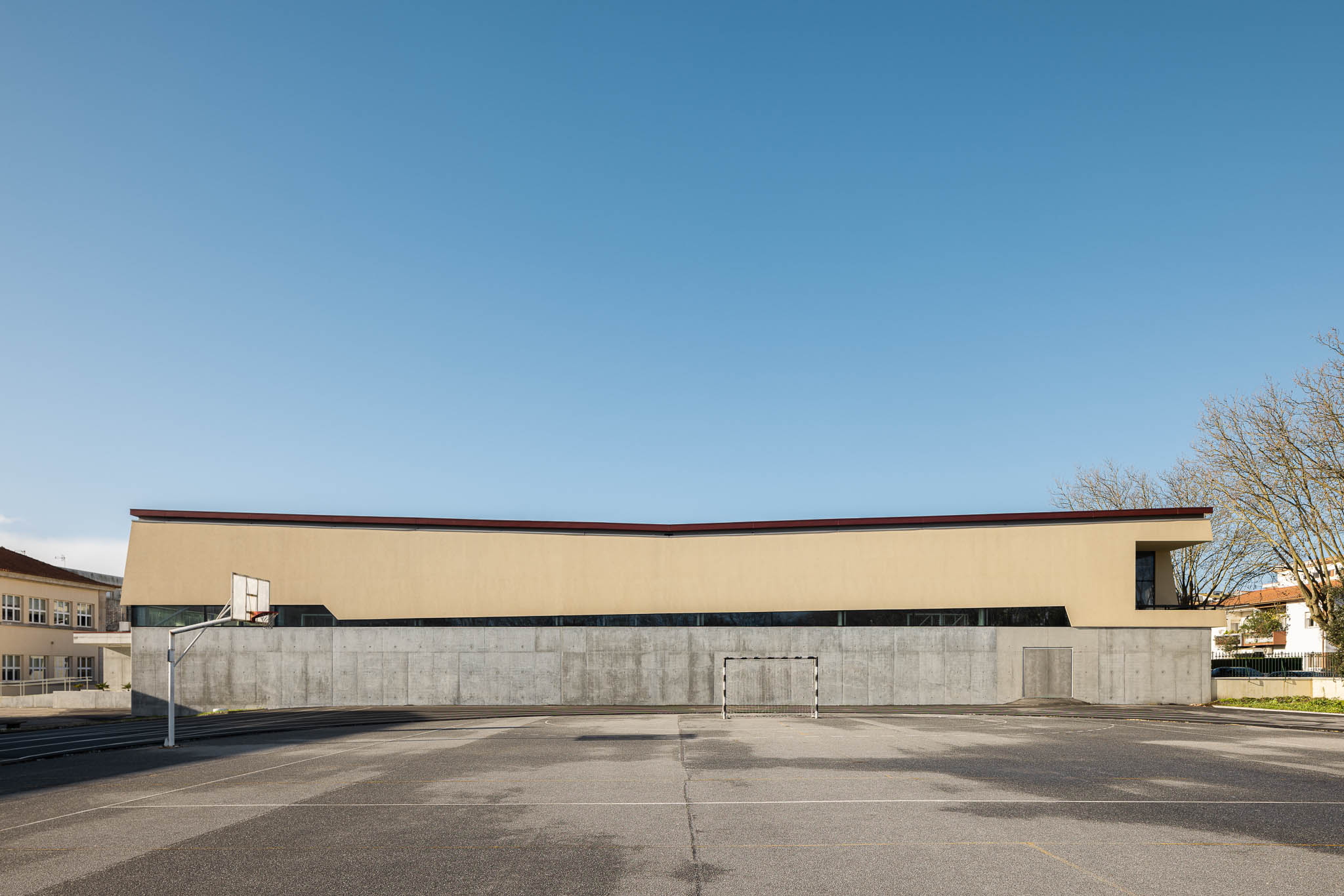 Fachada: Edifício com paredes em betão e estuque amarelo sob céu azul, campo de jogos em frente. — Pavilhão Desportivo da Escola Secundária Eça de Queirós arquitetura Oval em Póvoa de Varzim