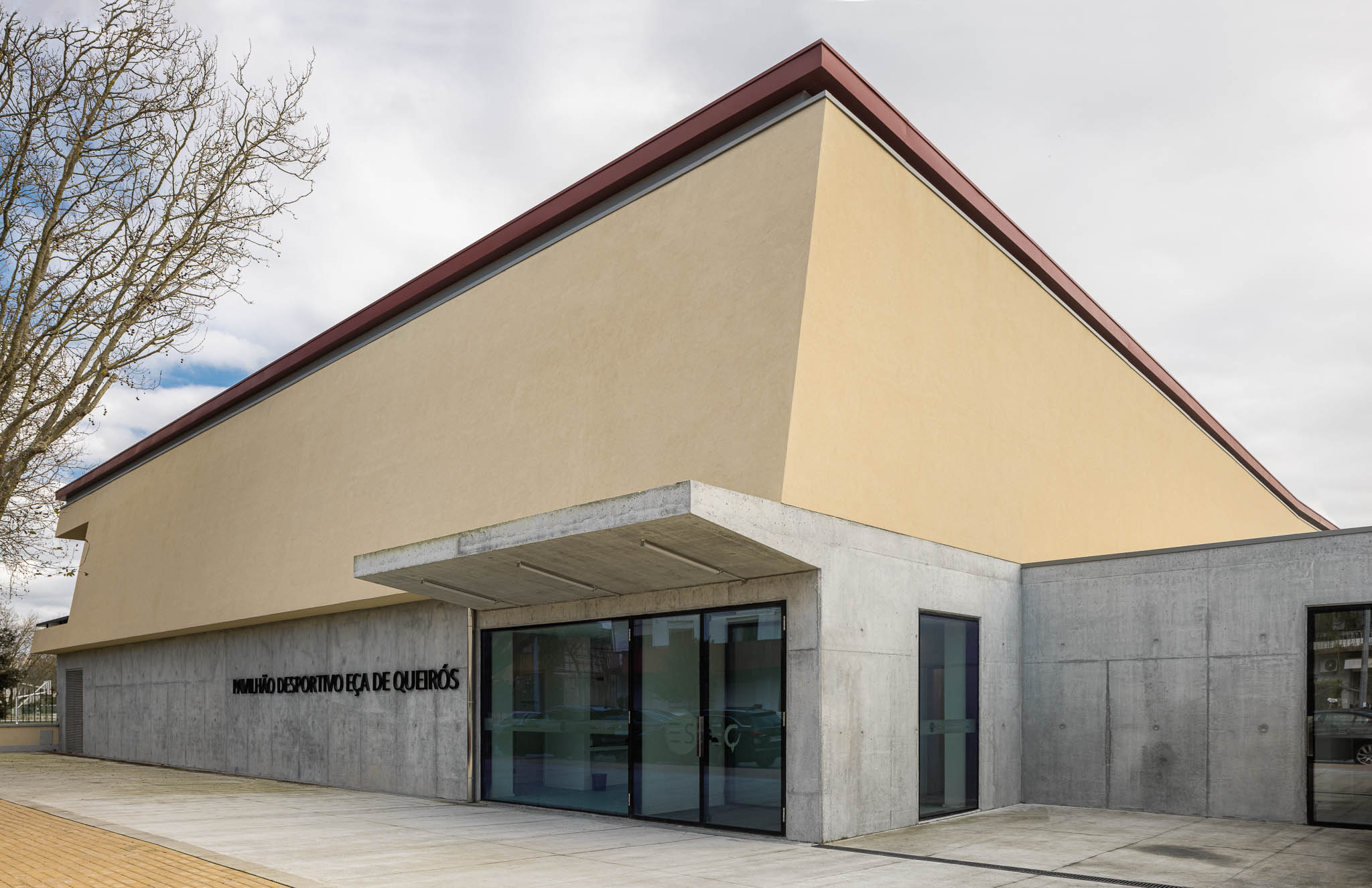 Fachada: Vista angular do pavilhão com paredes em betão e estuque amarelo, entrada envidraçada. — Pavilhão Desportivo da Escola Secundária Eça de Queirós arquitetura Oval em Póvoa de Varzim