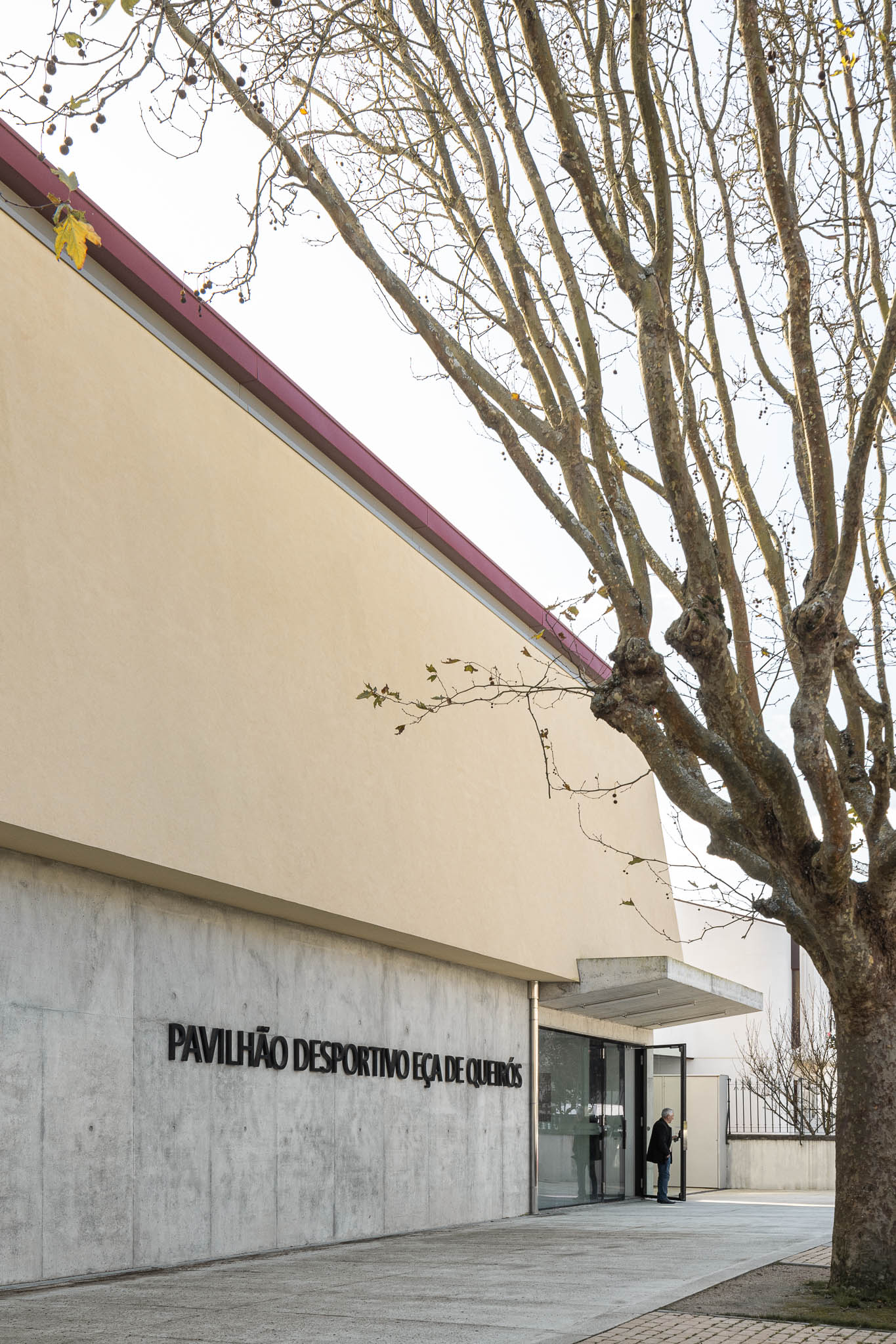 Fachada: Entrada do pavilhão com nome gravado em betão, árvore sem folhas em primeiro plano. — Pavilhão Desportivo da Escola Secundária Eça de Queirós arquitetura Oval em Póvoa de Varzim