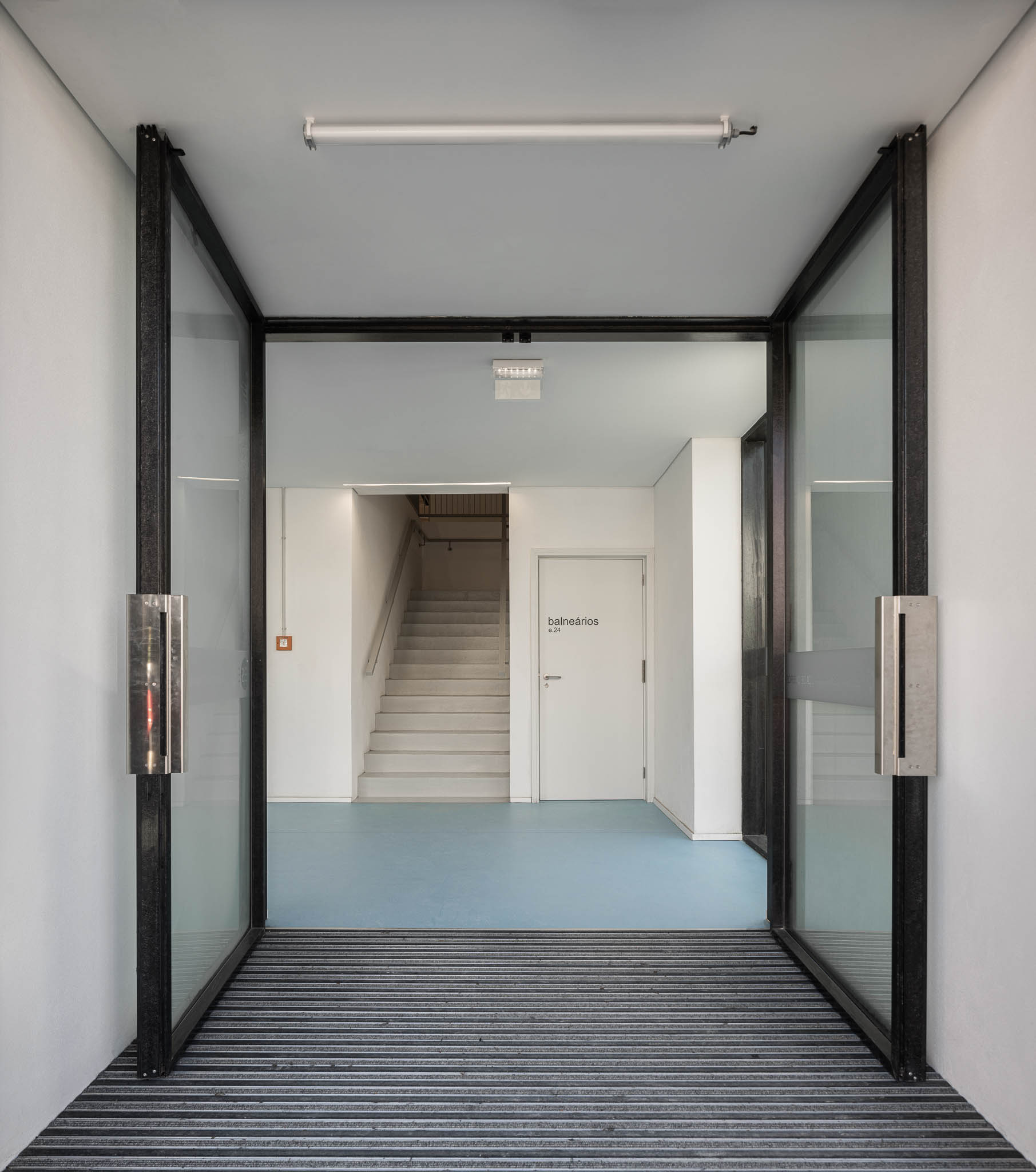 Entrada: Porta de vidro dupla aberta para um corredor com pavimento azul e escadas. — Pavilhão Desportivo da Escola Secundária Eça de Queirós arquitetura Oval em Póvoa de Varzim