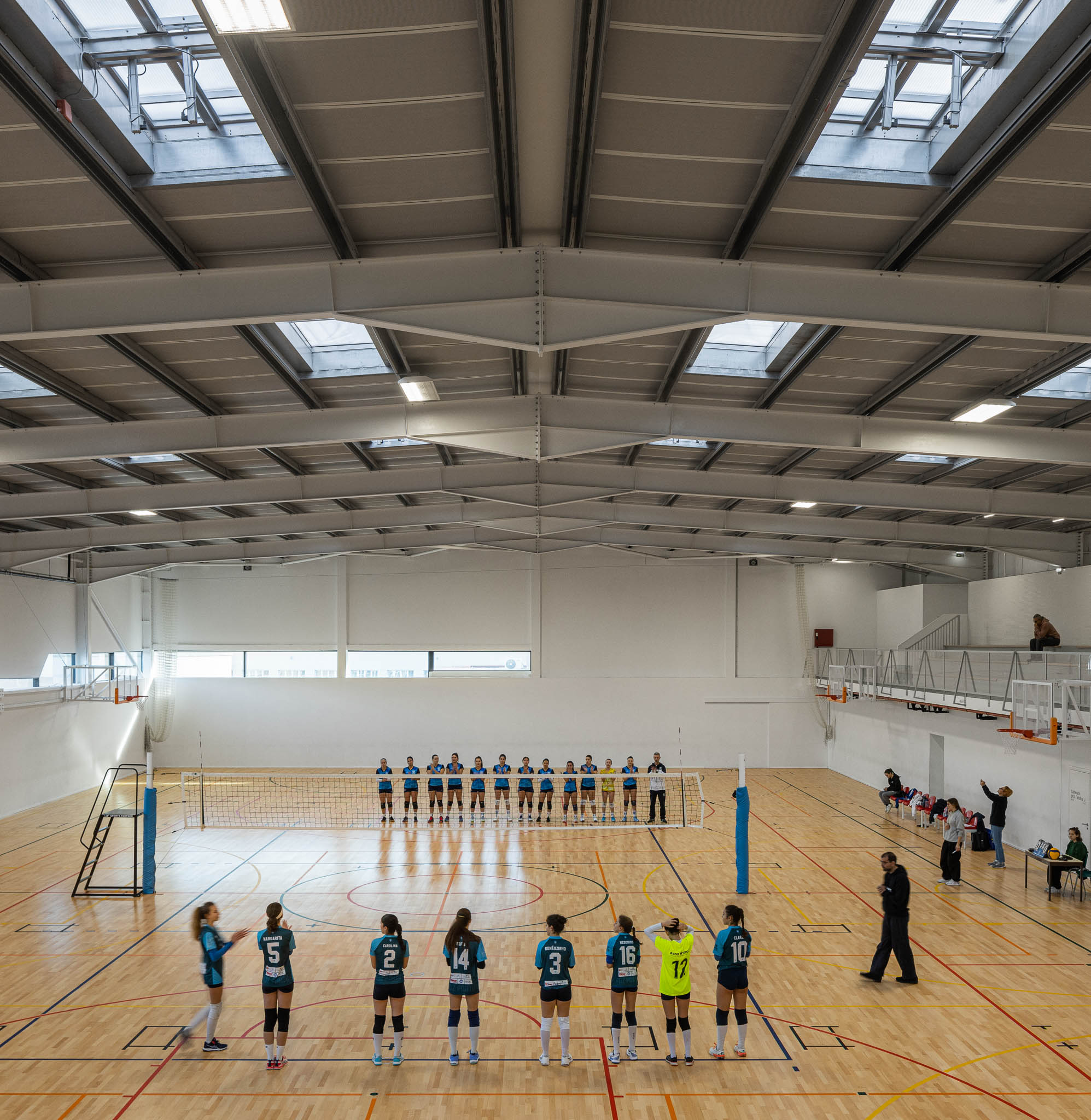 Pavilhão: Vista superior do campo de basquetebol com jogadores em ação, estrutura metálica no teto. — Pavilhão Desportivo da Escola Secundária Eça de Queirós arquitetura Oval em Póvoa de Varzim