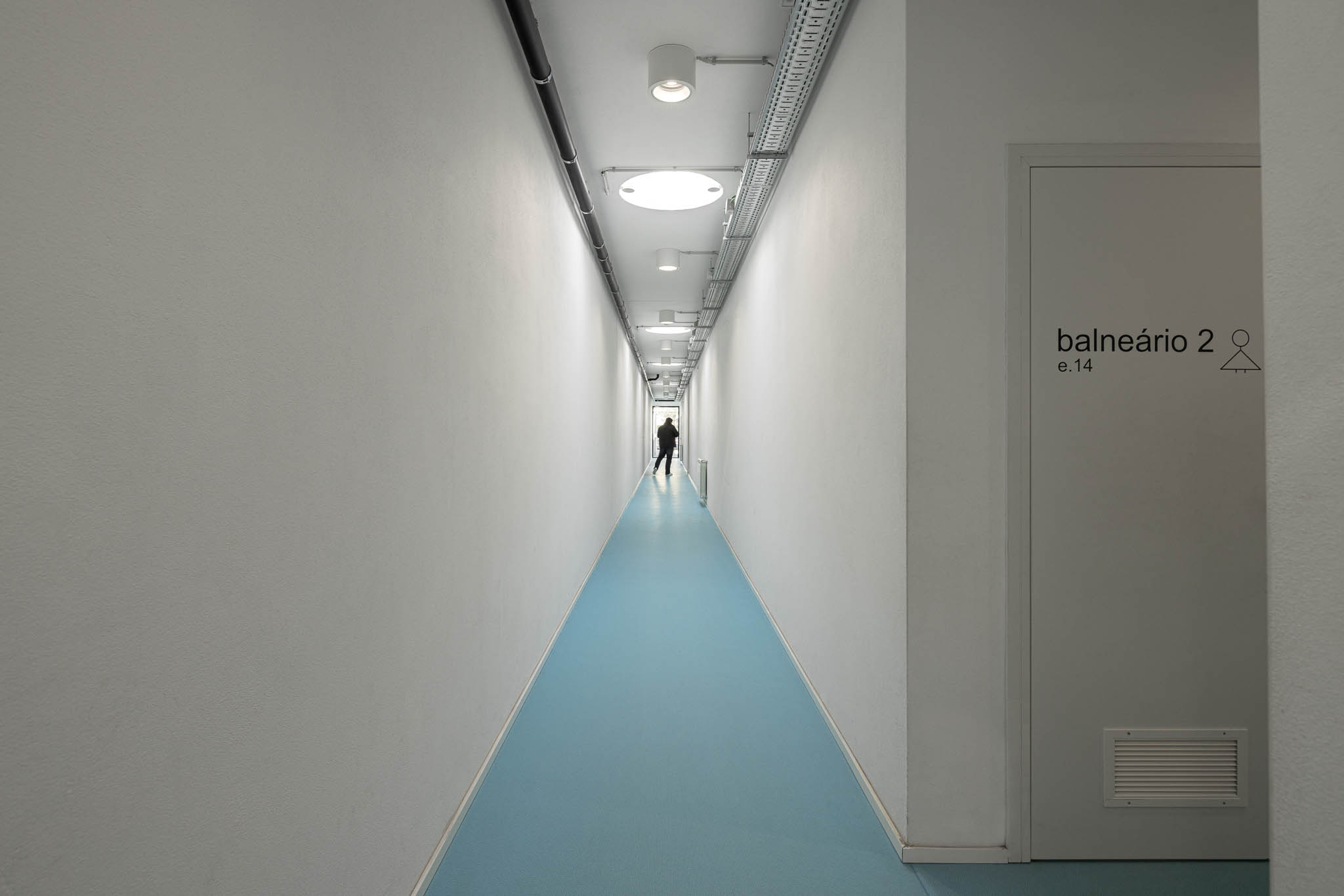 Corredor: Corredor com candeeiros cilíndricos e cablagem aparente no teto branco. — Pavilhão Desportivo da Escola Secundária Eça de Queirós arquitetura Oval em Póvoa de Varzim