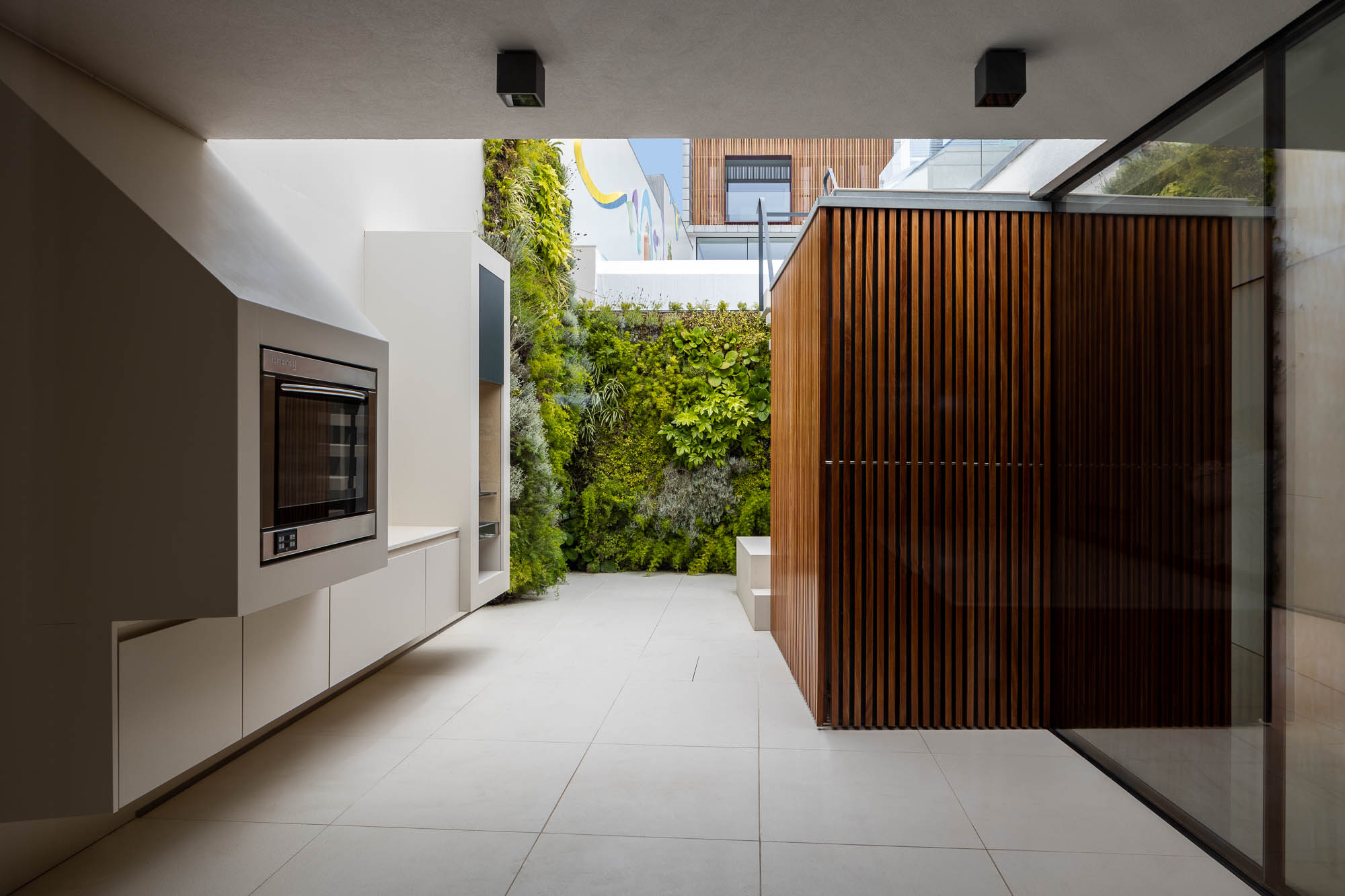Pátio: Cozinha exterior com armários brancos, churrasqueira e parede verde. Pavimento cerâmico claro e revestimento em madeira. — Casa Costa Nova arquitetura Espaço Objecto em Ílhavo