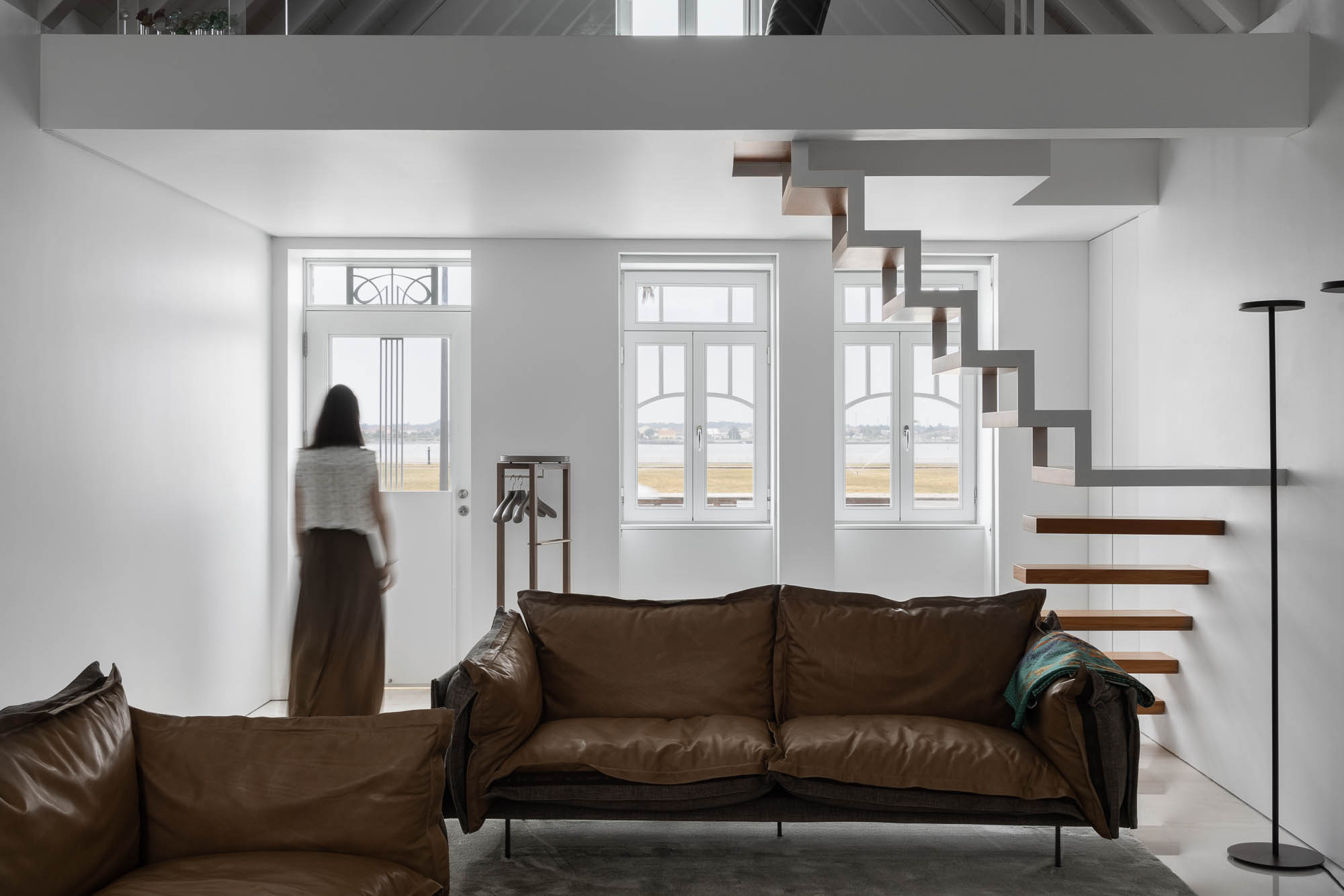 Sala de estar: Sofá de couro castanho, escadas flutuantes e vista para a ria através das janelas. Decoração minimalista e luminosa. — Casa Costa Nova arquitetura Espaço Objecto em Ílhavo