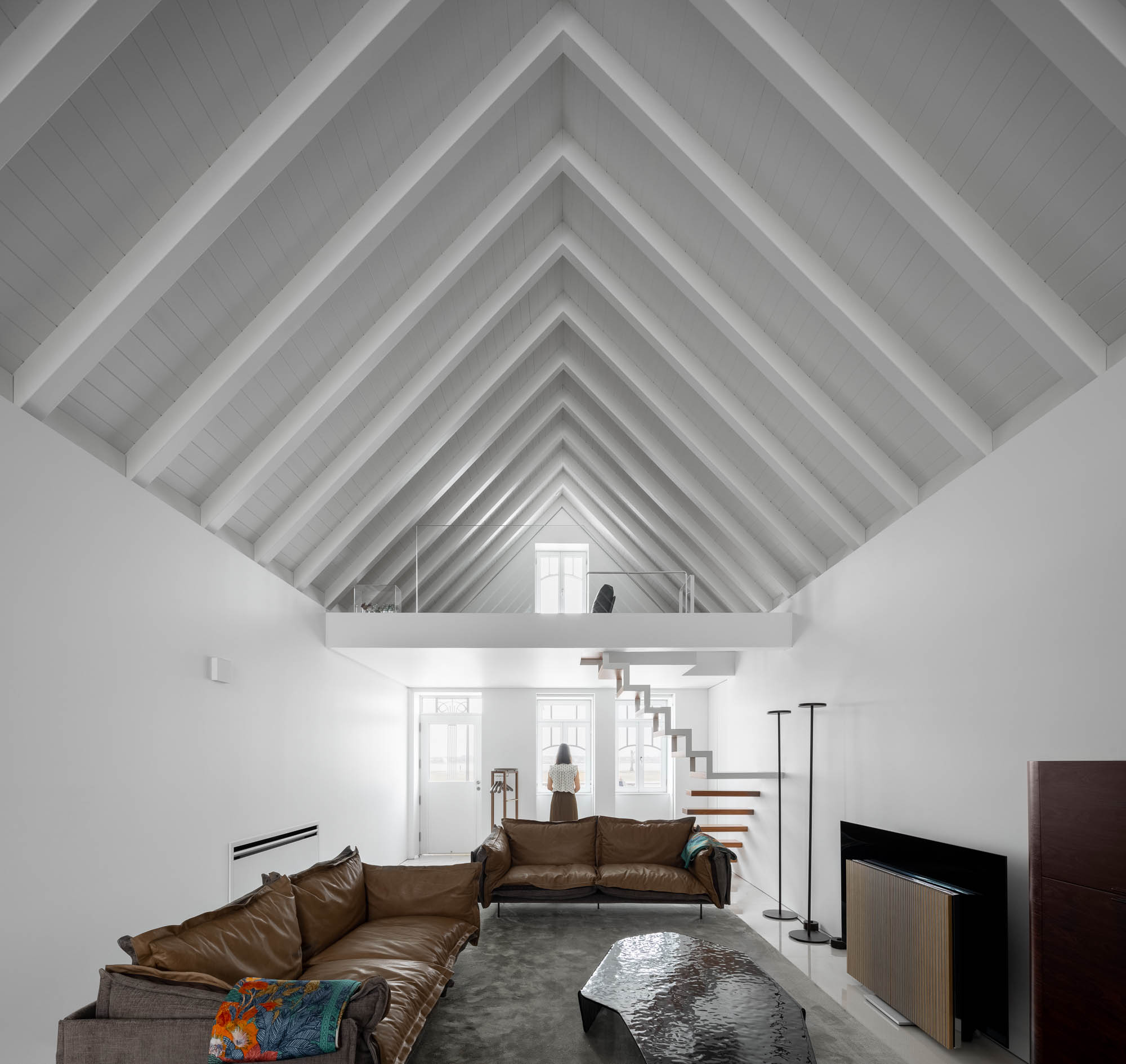 Sala de estar: Teto inclinado com vigas de madeira, sofá de couro, alcatifa cinzenta e escadas para mezanino. Espaço amplo e luminoso. — Casa Costa Nova arquitetura Espaço Objecto em Ílhavo