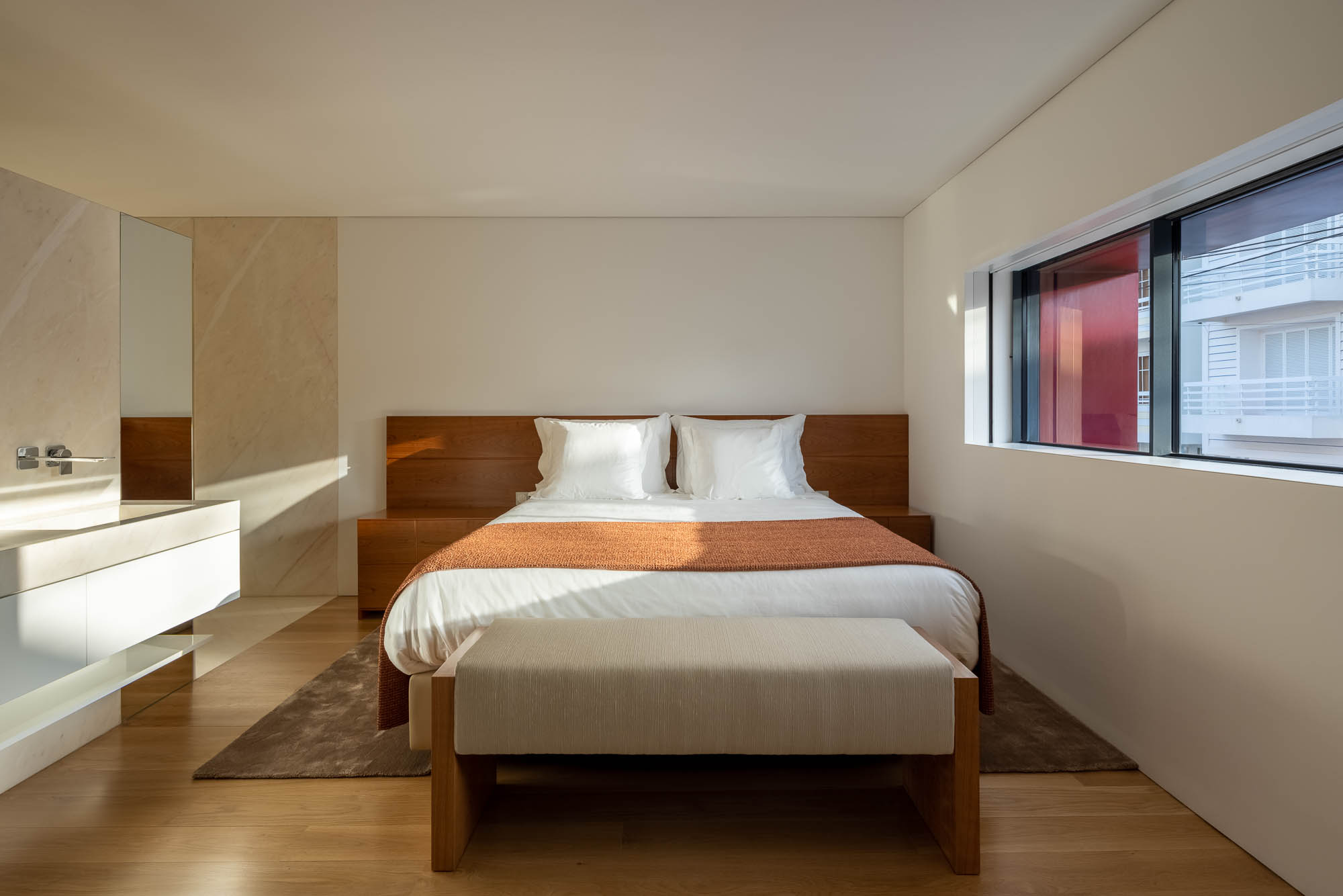 Quarto: Cama com roupa de cama cinzenta, casa de banho integrada com duche em mármore e lavatório em madeira escura. — Casa Costa Nova arquitetura Espaço Objecto em Ílhavo