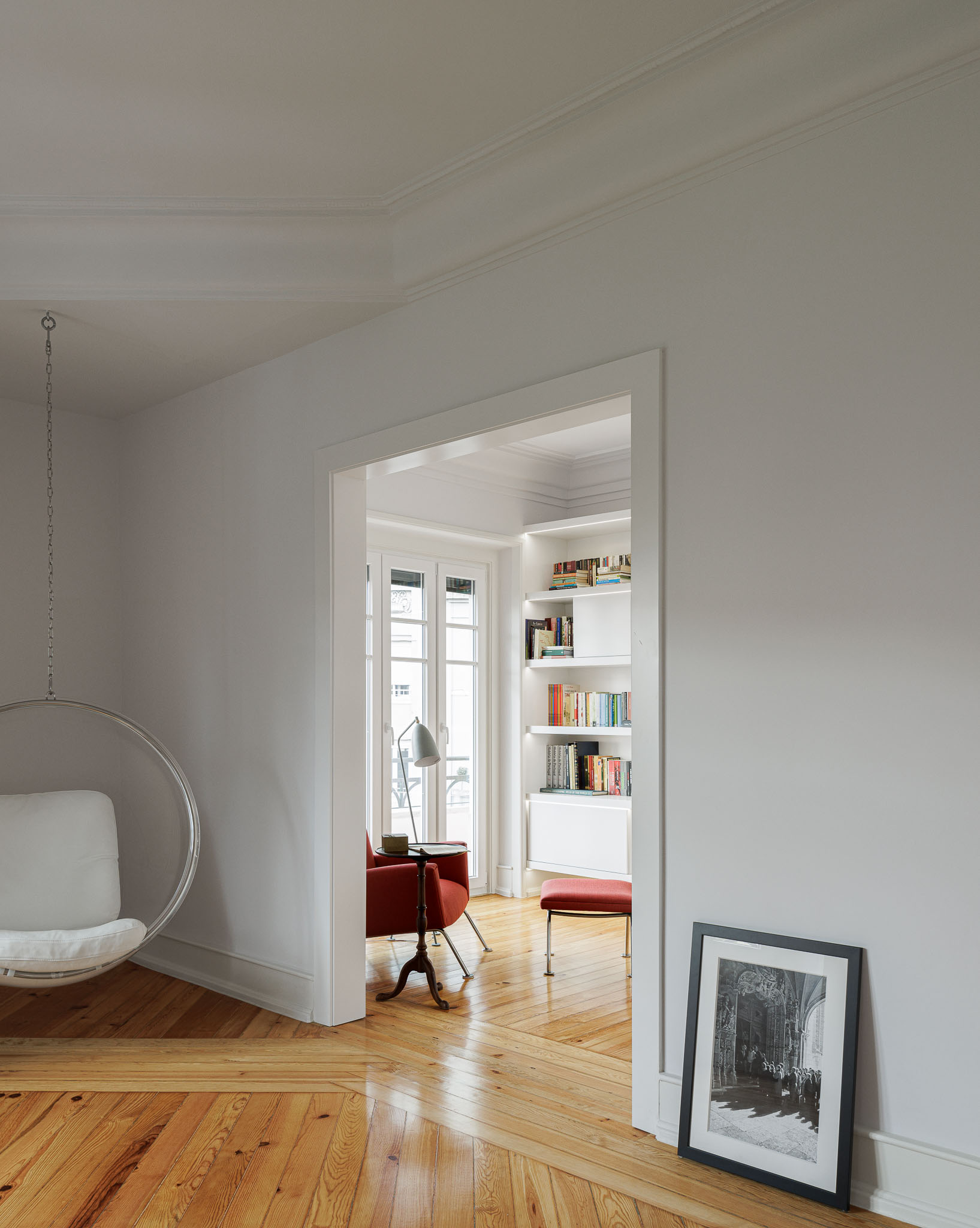 Escritório: Cadeira suspensa, vista para a varanda com estantes brancas e cadeira vermelha. Pavimento em madeira. — Apartamento AN arquitetura NMDARQ – Nuno Miguel Dias, Arquitecto em Lisboa