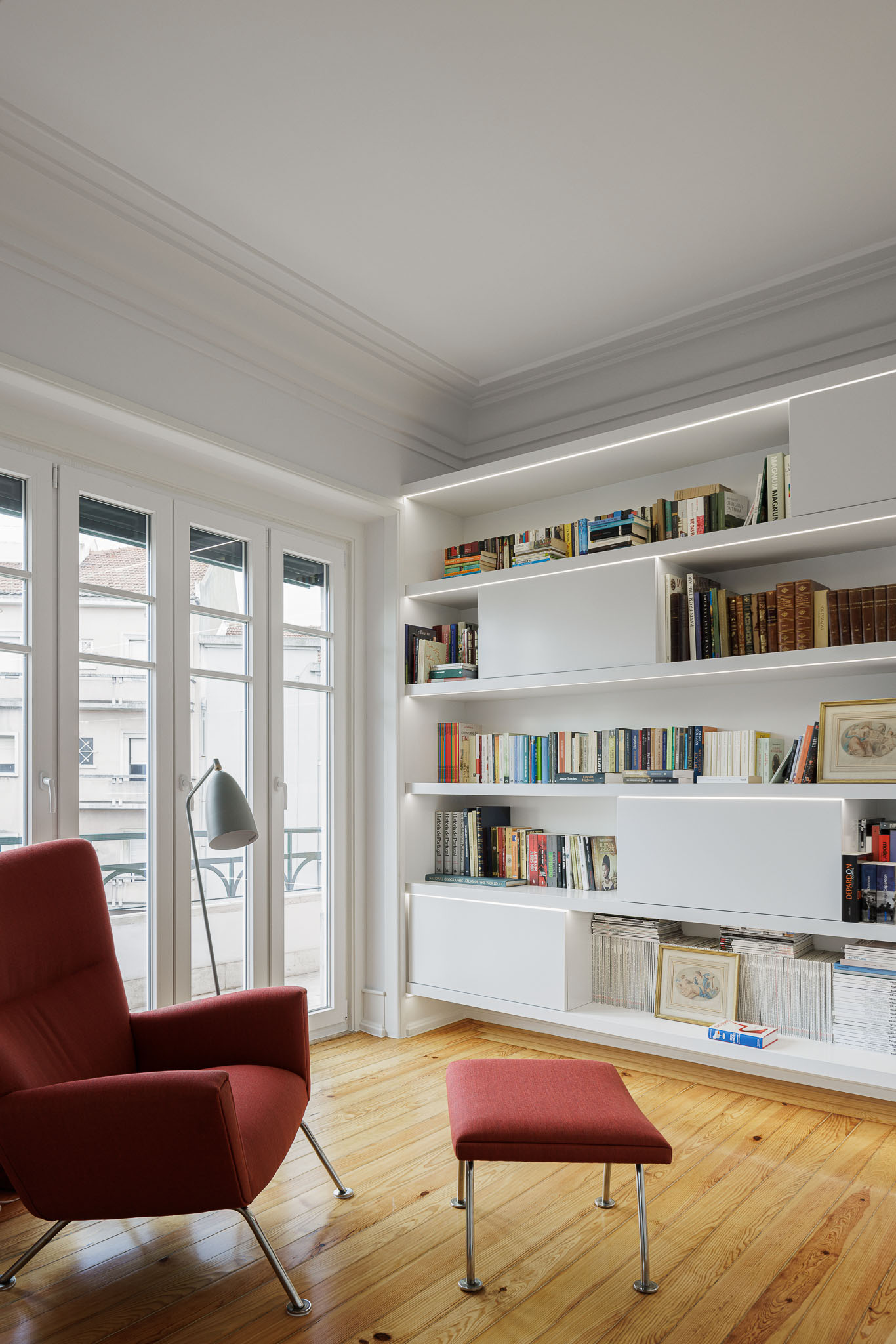 Escritório: Estantes brancas iluminadas, cadeira e pufe vermelhos, pavimento em madeira e acesso à varanda. — Apartamento AN arquitetura NMDARQ – Nuno Miguel Dias, Arquitecto em Lisboa