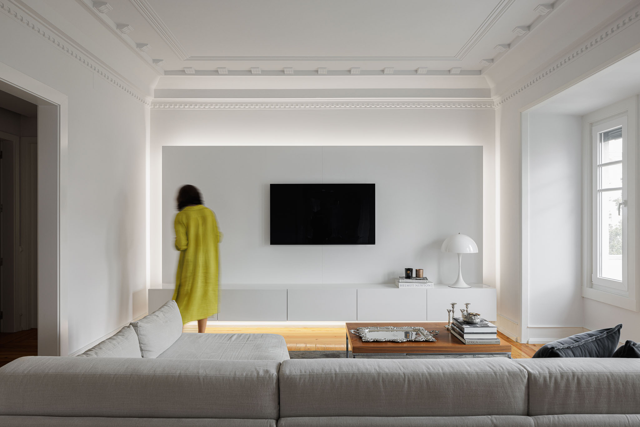 Sala de estar: Painel branco iluminado com televisão, sofá cinzento e mesa de centro com livros e objetos decorativos. — Apartamento AN arquitetura NMDARQ – Nuno Miguel Dias, Arquitecto em Lisboa