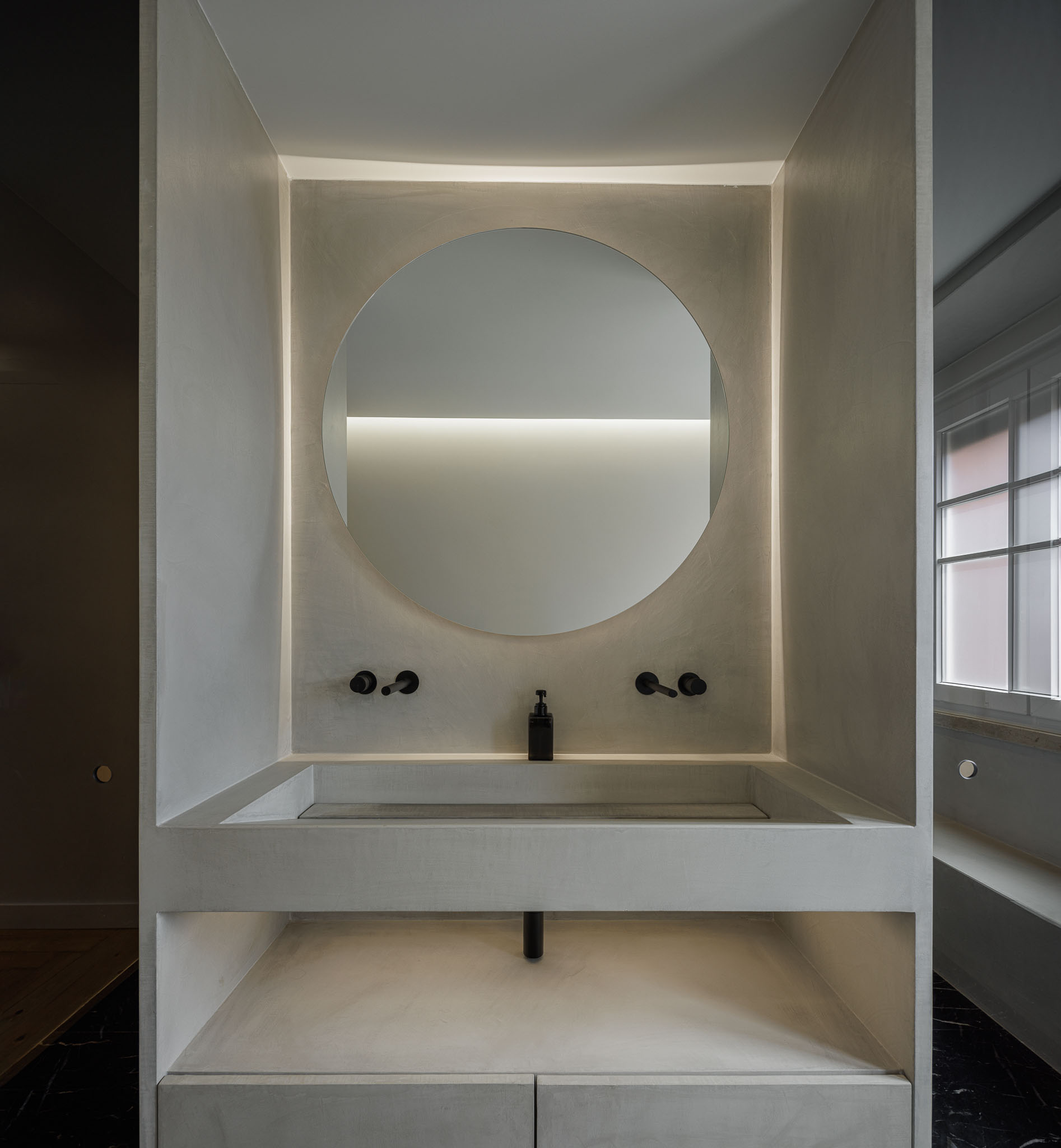Casa de banho: Lavatório em betão aparente com espelho redondo e iluminação embutida. — Apartamento AN arquitetura NMDARQ – Nuno Miguel Dias, Arquitecto em Lisboa