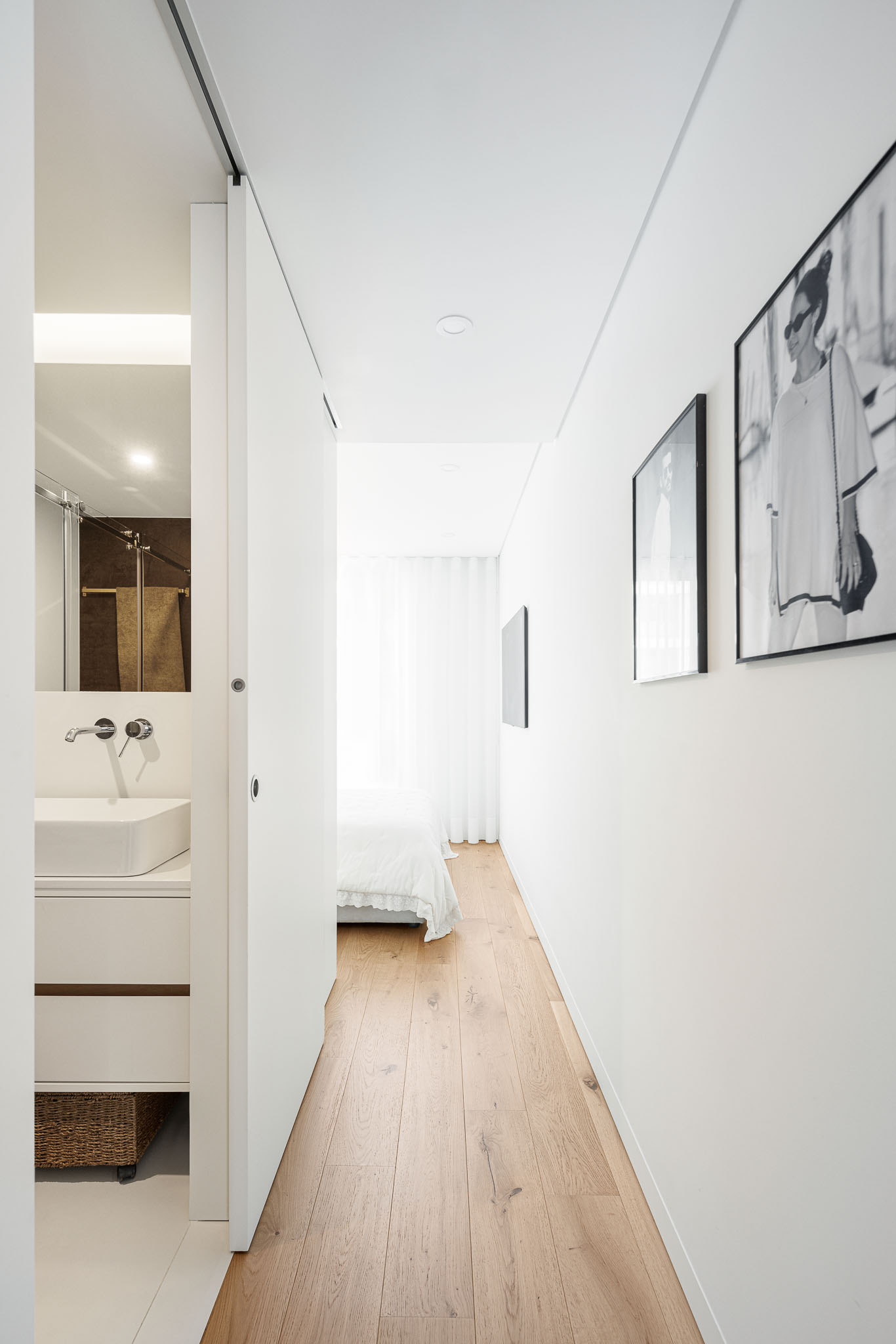 Casa de banho: Lavatório branco moderno com espelho, porta de correr e vista para o quarto com cama. — 5ª Avenida arquitetura Mário Alves Arquitecto em Aveiro