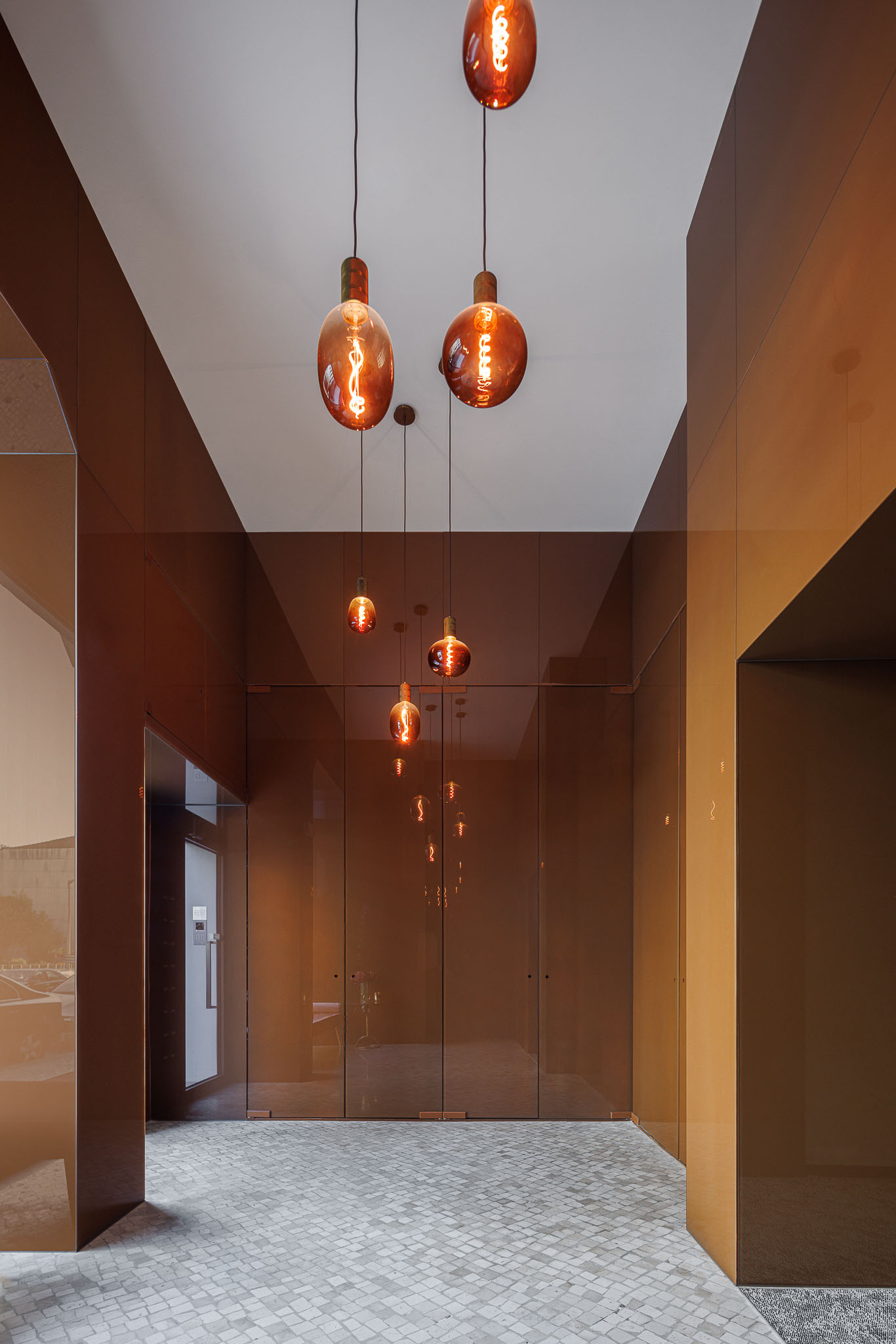Hall de entrada: Corredor com paredes revestidas a espelho bronze, candeeiros suspensos e pavimento em calçada. — 5ª Avenida arquitetura Mário Alves Arquitecto em Aveiro