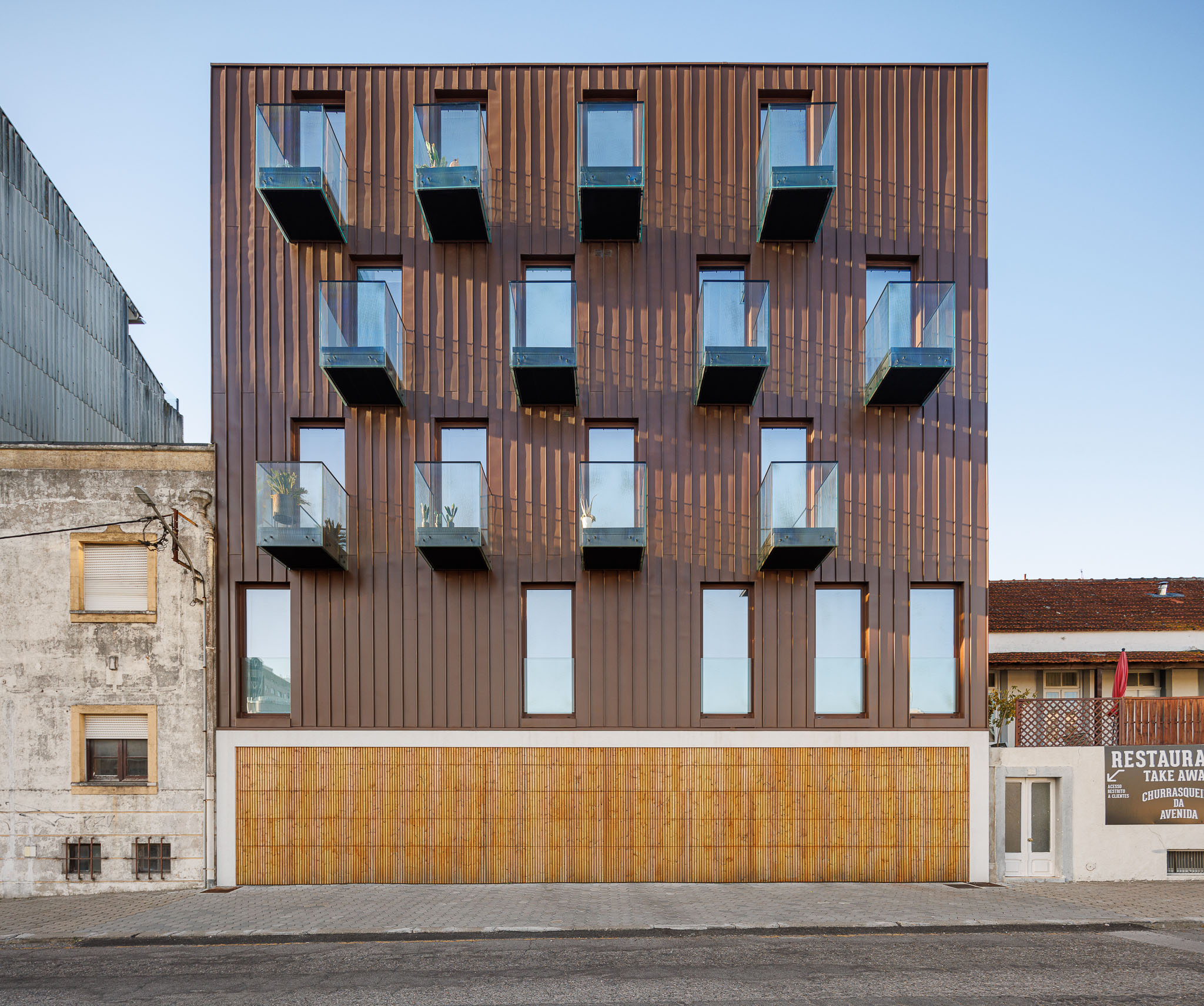 Fachada: Vista frontal do edifício com fachada em cobre, varandas de vidro e base em madeira ripada. — 5ª Avenida arquitetura Mário Alves Arquitecto em Aveiro