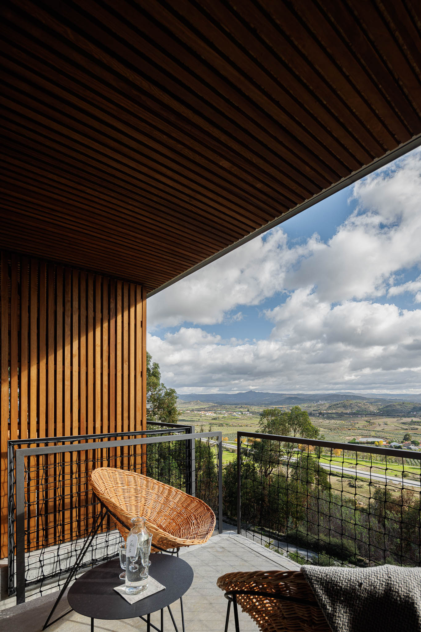 Varanda: Cadeiras de vime e mesa preta, com vista para a paisagem. Teto em madeira ripada. — The Vagar – Country House arquitetura David Bilo Arquitectos e Filipe Pina Arquitectura em Belmonte