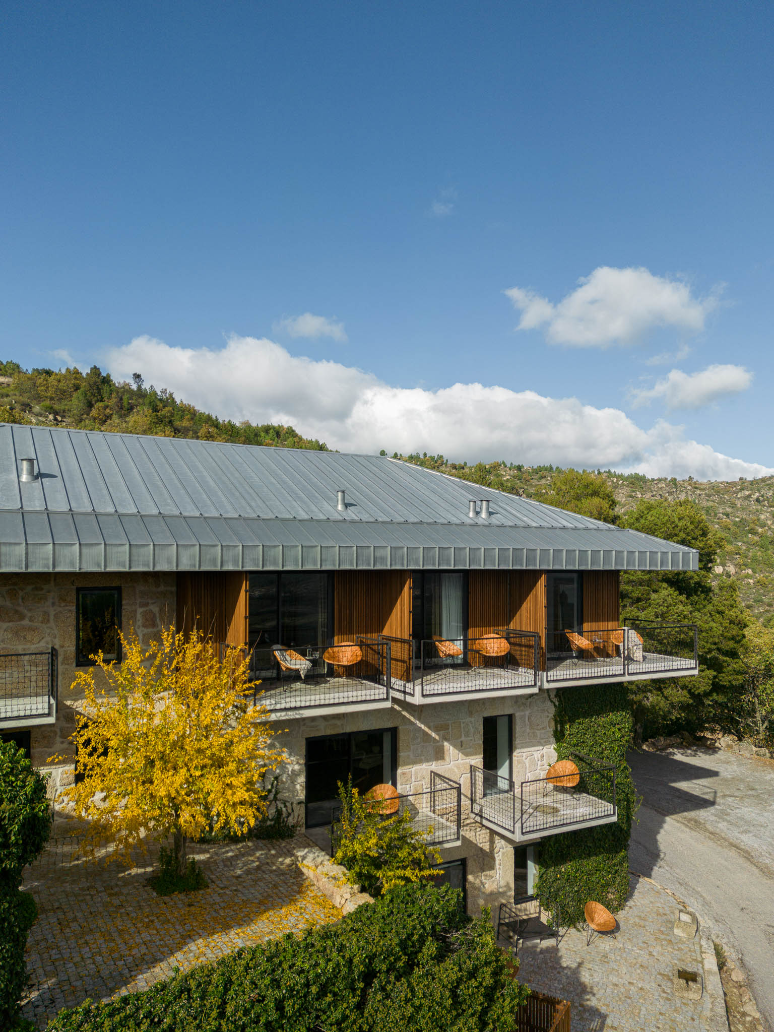 Exterior: Vista geral da casa de campo com revestimento em pedra e madeira, varandas com cadeiras de vime. — The Vagar – Country House arquitetura David Bilo Arquitectos e Filipe Pina Arquitectura em Belmonte