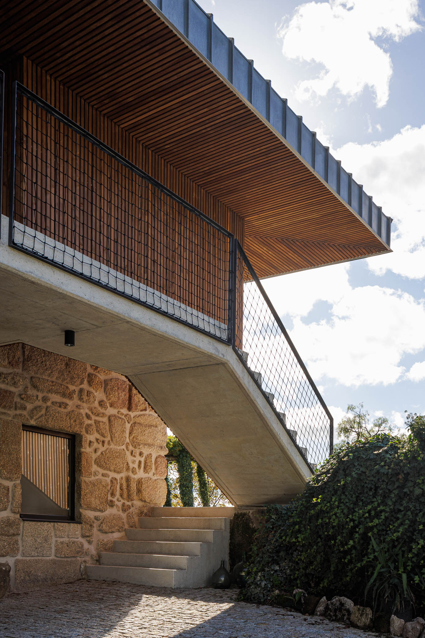 Exterior: Escadas de acesso ao primeiro andar em betão, com guarda em rede metálica preta. Revestimento em madeira. — The Vagar – Country House arquitetura David Bilo Arquitectos e Filipe Pina Arquitectura em Belmonte