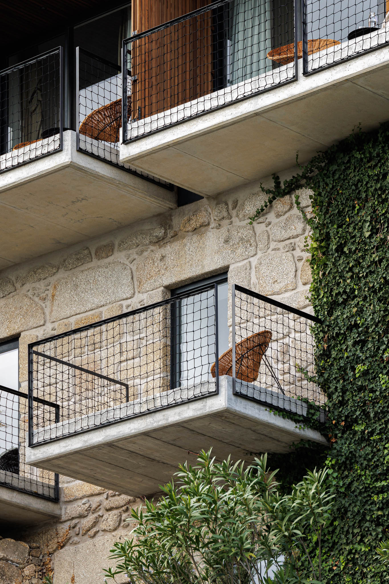 Exterior: Fachada em pedra com varandas em betão e guarda em rede metálica preta. Cadeiras de vime nas varandas. — The Vagar – Country House arquitetura David Bilo Arquitectos e Filipe Pina Arquitectura em Belmonte