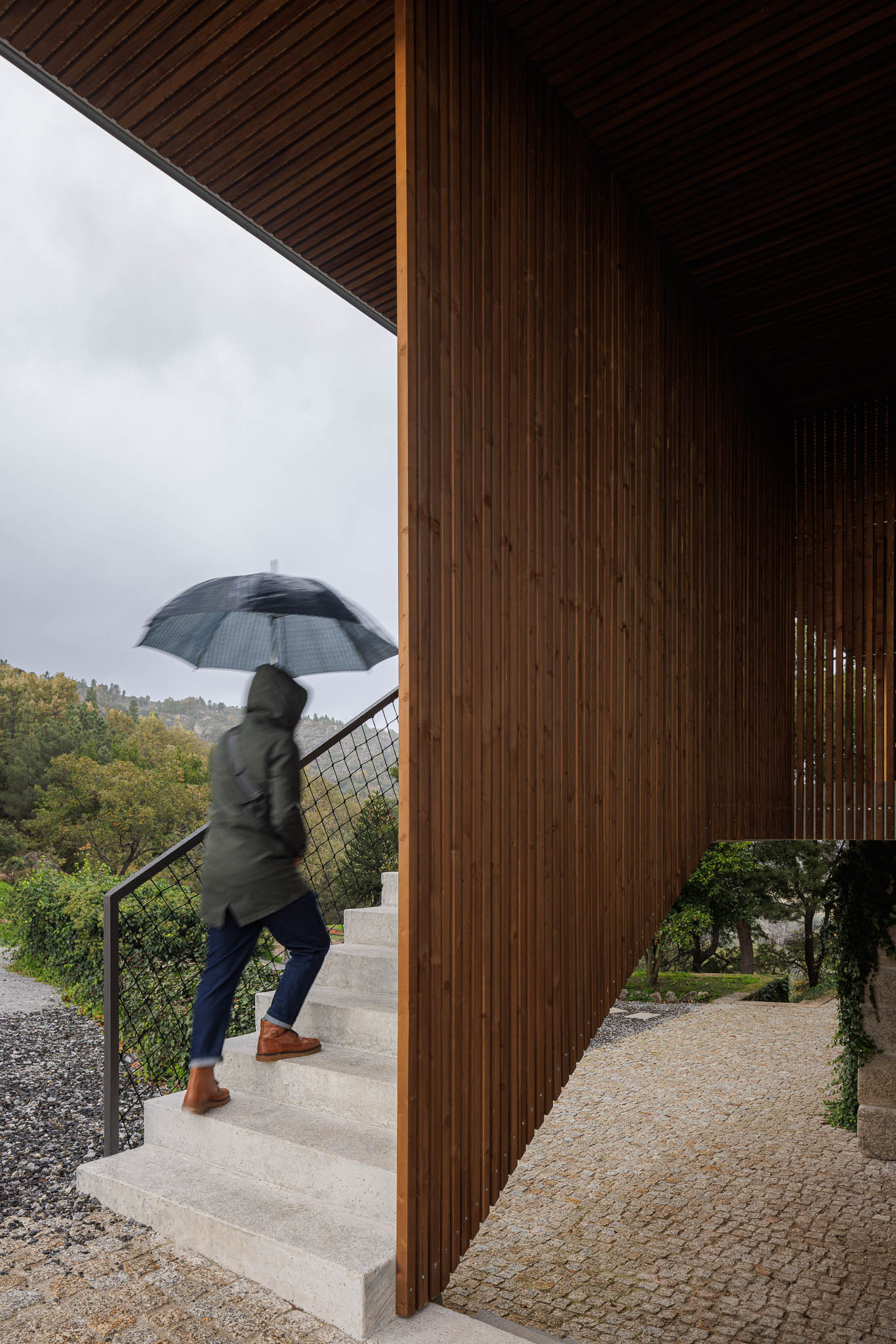 Exterior: Pessoa a subir as escadas de acesso em betão com guarda em rede metálica preta. Revestimento em madeira. — The Vagar – Country House arquitetura David Bilo Arquitectos e Filipe Pina Arquitectura em Belmonte