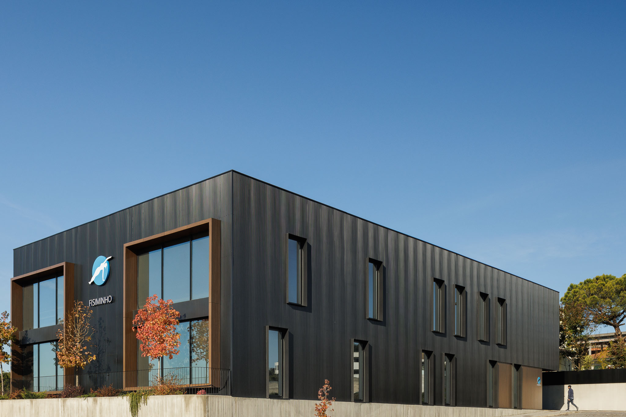 Fachada: Perspectiva angular do edifício com revestimento metálico preto, janelas e paisagismo. — Clínica de fisioterapia - Fisiminho arquitetura L2C Arquitetura em Braga