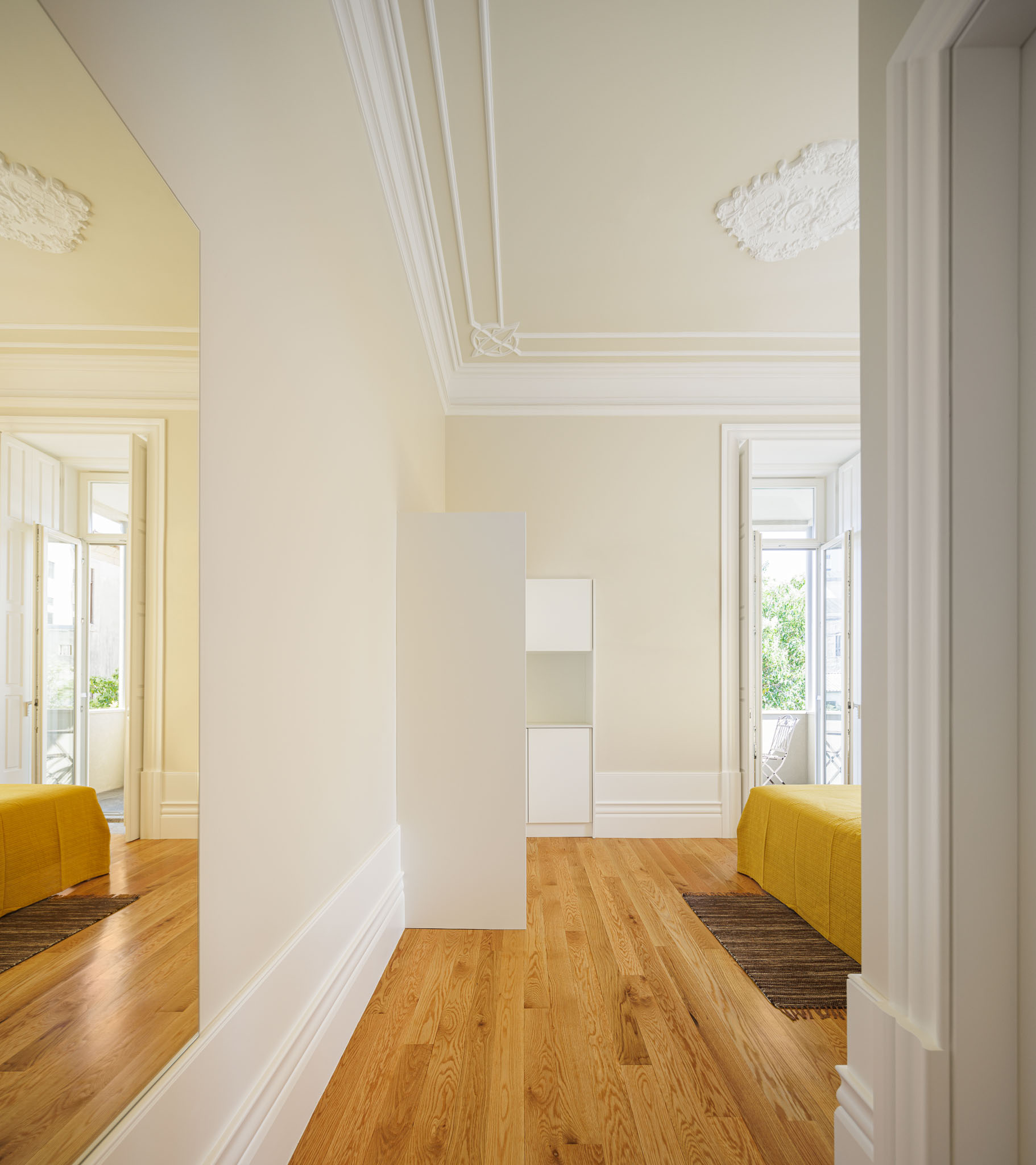 Quarto: Espelho reflectindo cama amarela e pavimento de madeira, com detalhes clássicos no tecto e rodapés brancos. — Casa Kala arquitetura Ren Ito em Porto