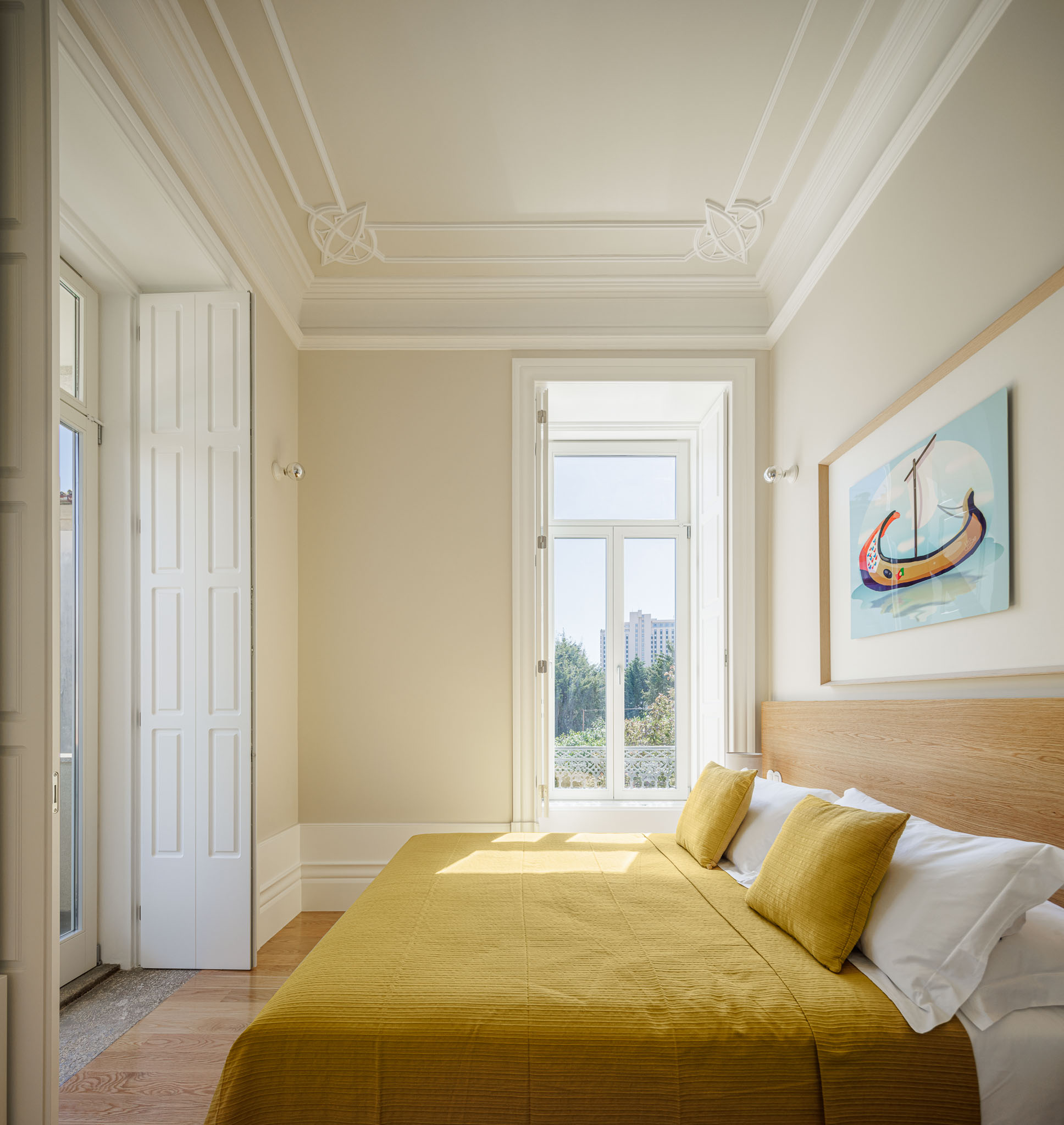 Quarto: Cama com roupa de cama amarela, cabeceira de madeira, janela com portadas brancas e tecto com molduras clássicas. — Casa Kala arquitetura Ren Ito em Porto