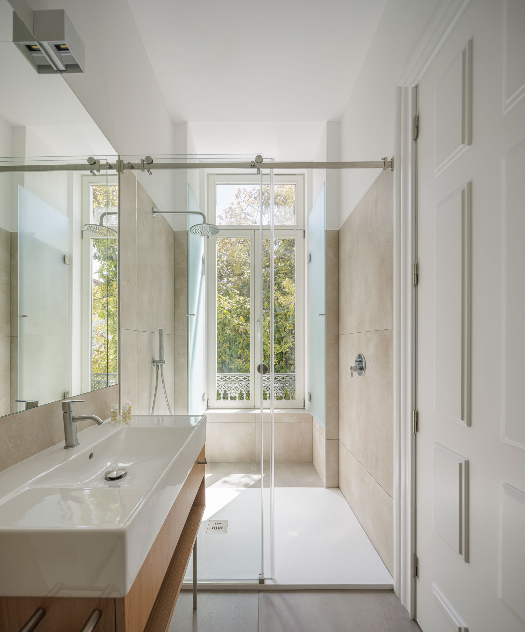 Casa de banho: Lavatório branco sobre móvel de madeira, duche com divisória de vidro e janela com vista para o exterior. — Casa Kala arquitetura Ren Ito em Porto