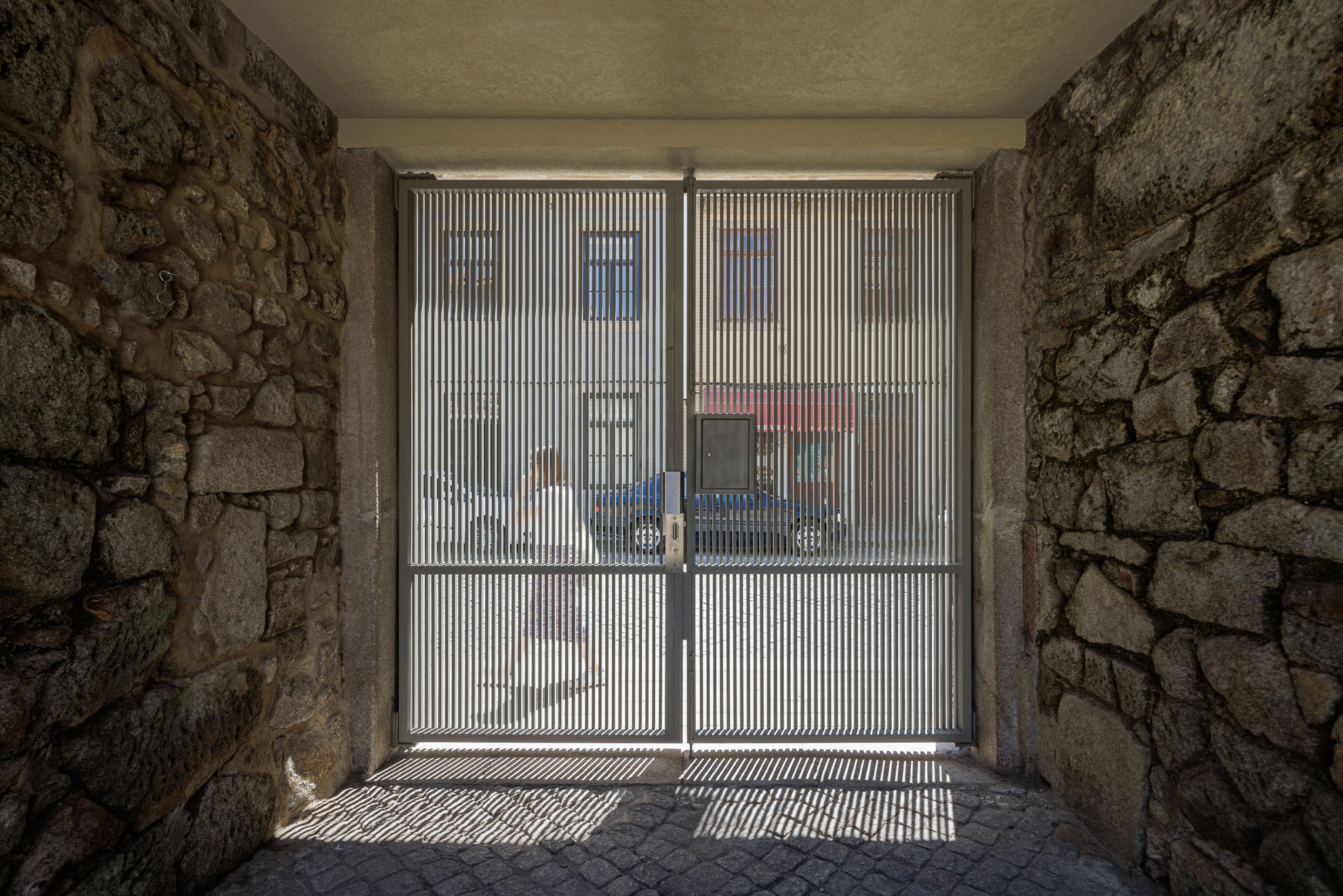 Exterior: Portão metálico com ripas verticais, paredes de pedra rústica e pavimento de calçada. — Casa Kala arquitetura Ren Ito em Porto