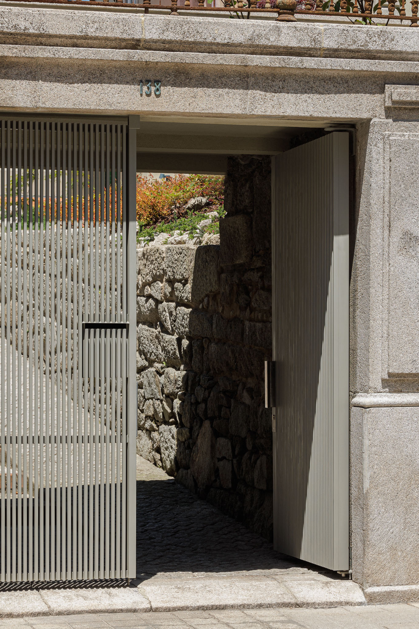 Exterior: Detalhe do portão metálico aberto, com paredes de pedra e vegetação ao fundo. — Casa Kala arquitetura Ren Ito em Porto