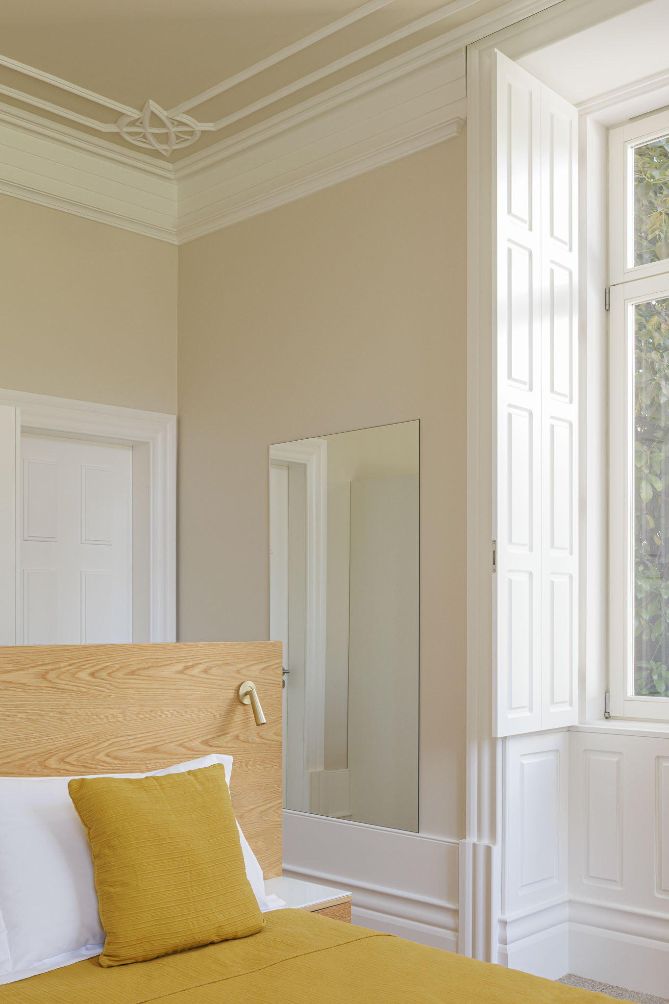 Quarto: Cama com roupa de cama amarela, espelho, janela com portadas brancas e tecto com molduras clássicas. — Casa Kala arquitetura Ren Ito em Porto