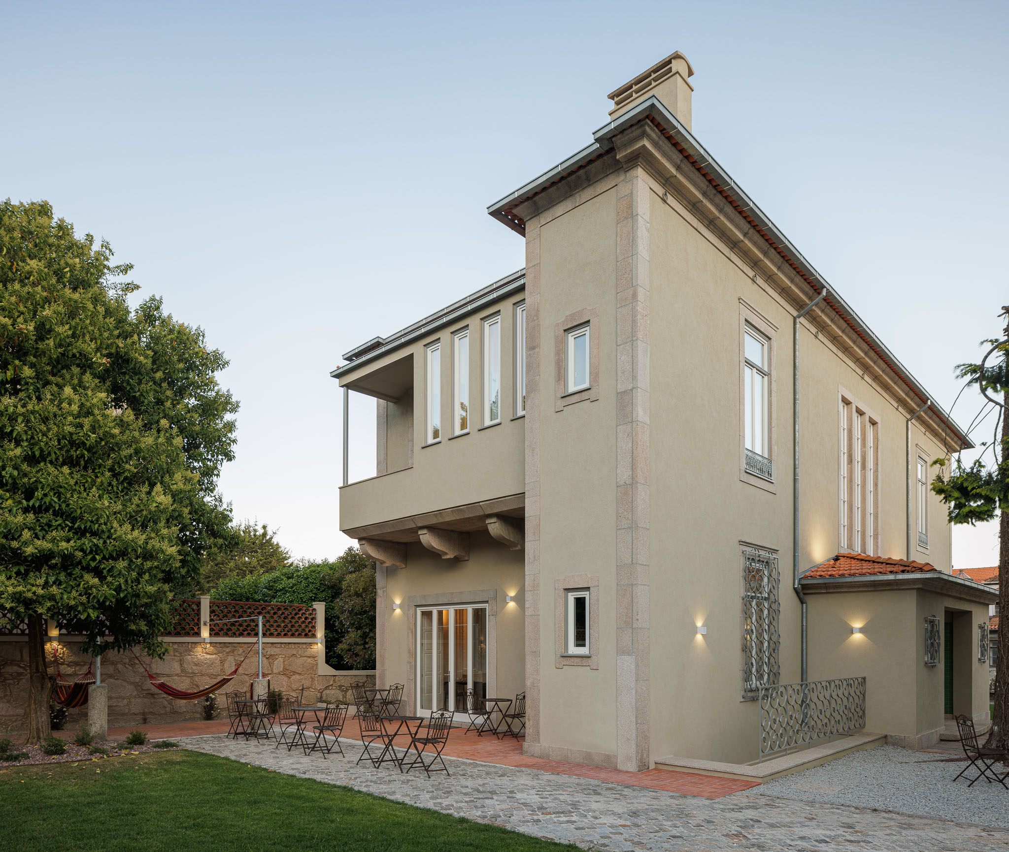 Exterior: Vista lateral da casa com jardim relvado, árvores e área de estar com cadeiras. — Casa Kala arquitetura Ren Ito em Porto