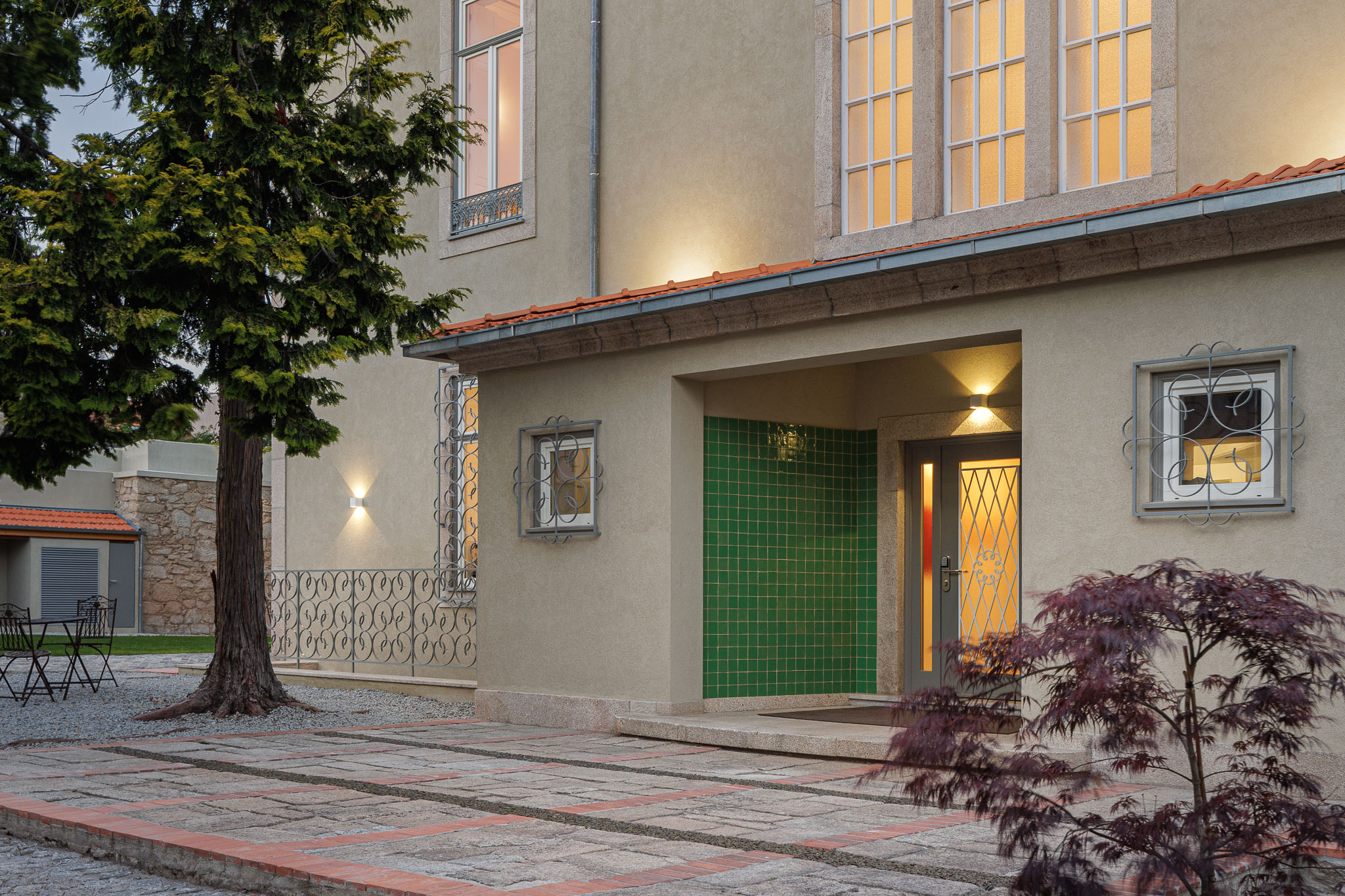 Exterior: Fachada da casa com revestimento bege, telhado vermelho, entrada com azulejos verdes e jardim com árvores. — Casa Kala arquitetura Ren Ito em Porto