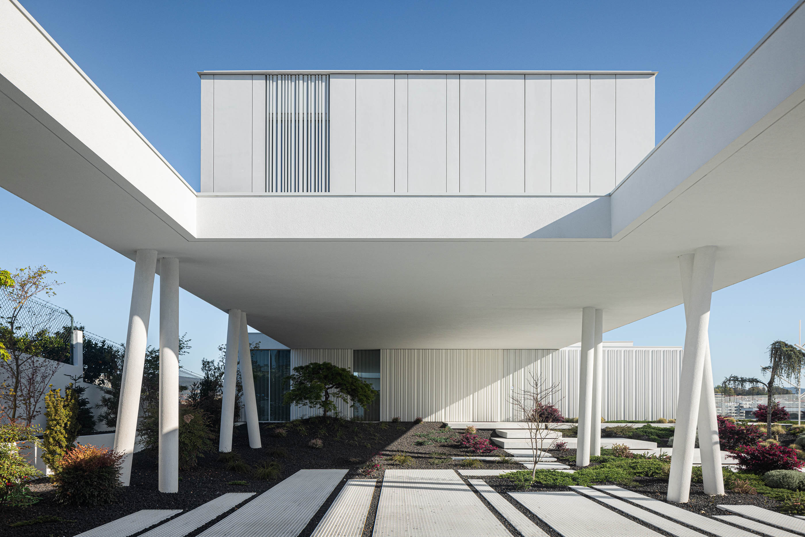 Jardim: Perspectiva inferior da moradia branca, com pilares inclinados e jardim minimalista com caminhos em grelha. — Riscowhite arquitetura Risco Singular em Barcelos