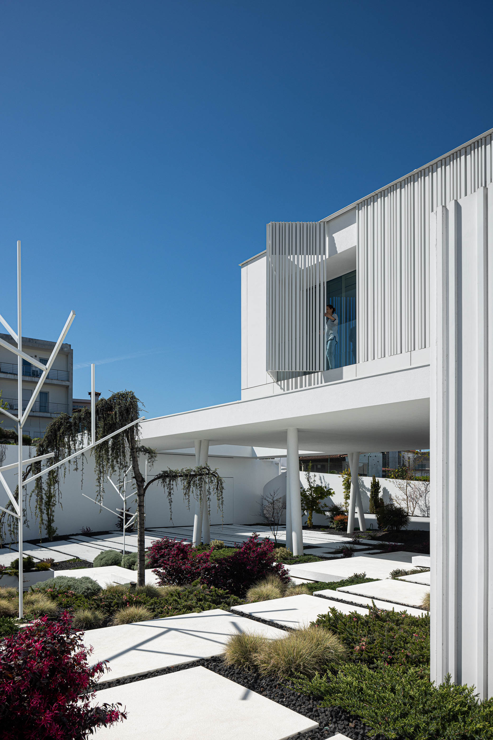 Jardim: Moradia branca com jardim minimalista, escada em caracol e estrutura metálica decorativa, com pessoa na janela. — Riscowhite arquitetura Risco Singular em Barcelos
